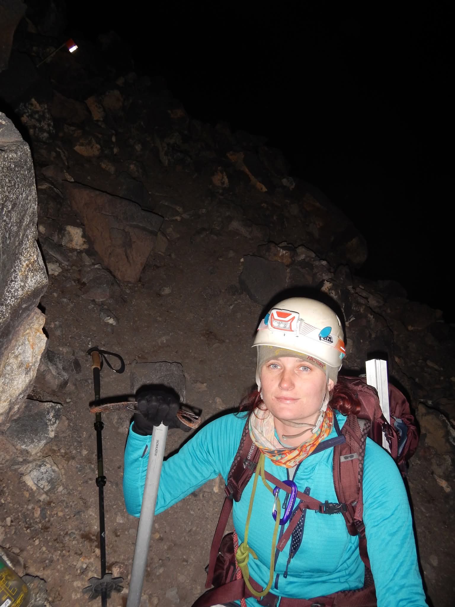Person wearing a helmet and backpack, sitting outdoors at night with rocky terrain around.