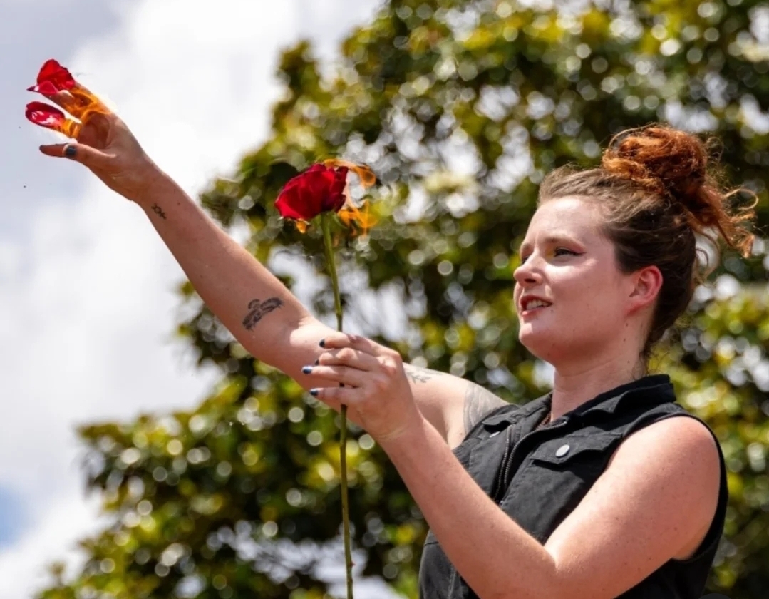 Young woman with red hair in a bun holds a rose and sprays water outdoors, with trees in the background.