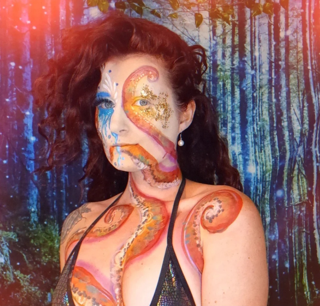 Woman with colorful face and body paint, standing in a forest background, smiling, wearing a black top with thin straps.