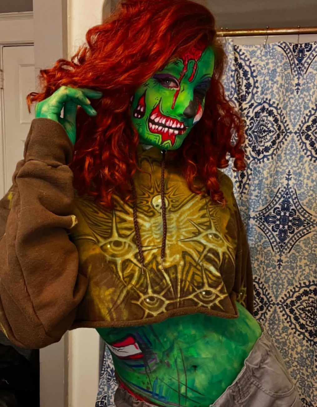 Person with green face paint and red curly hair, wearing a brown hoodie, standing indoors near a patterned curtain.