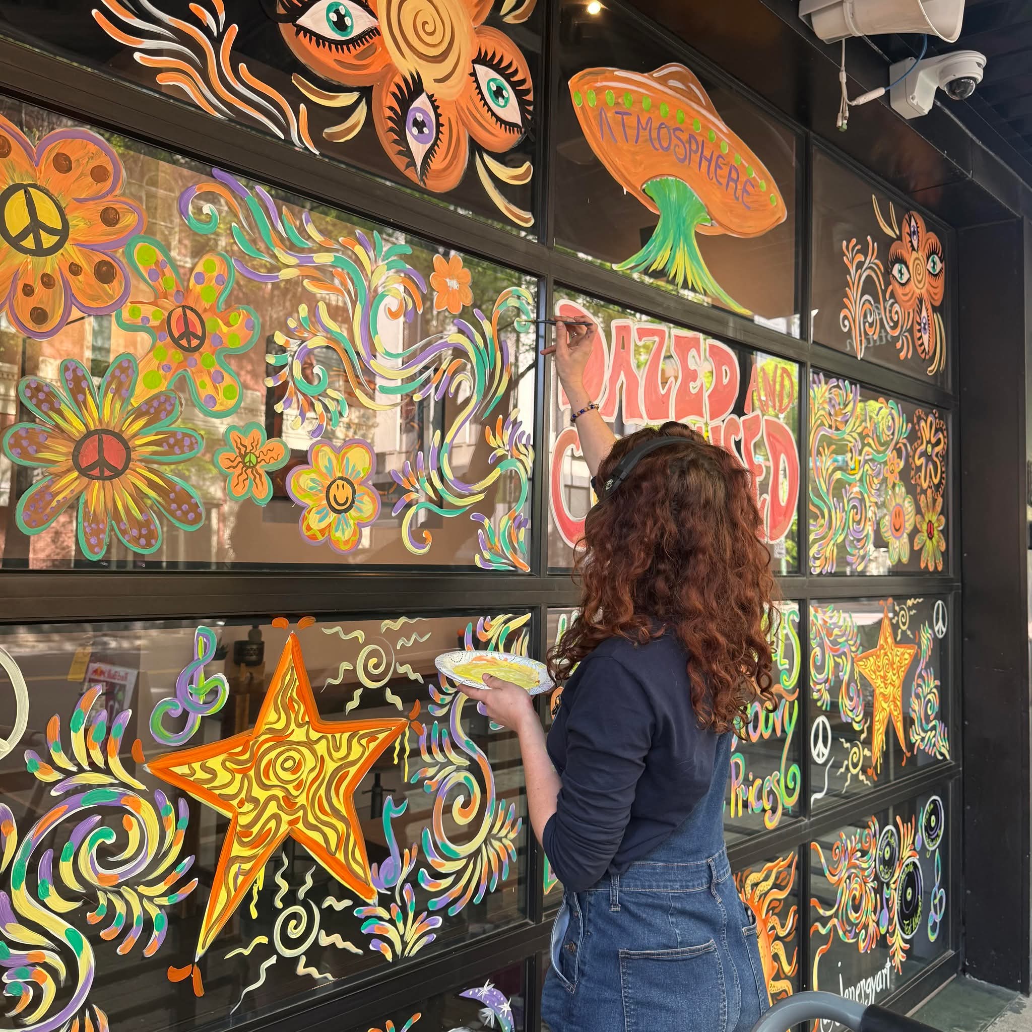 Woman painting colorful, psychedelic floral and star designs on a black wall with multiple panels.