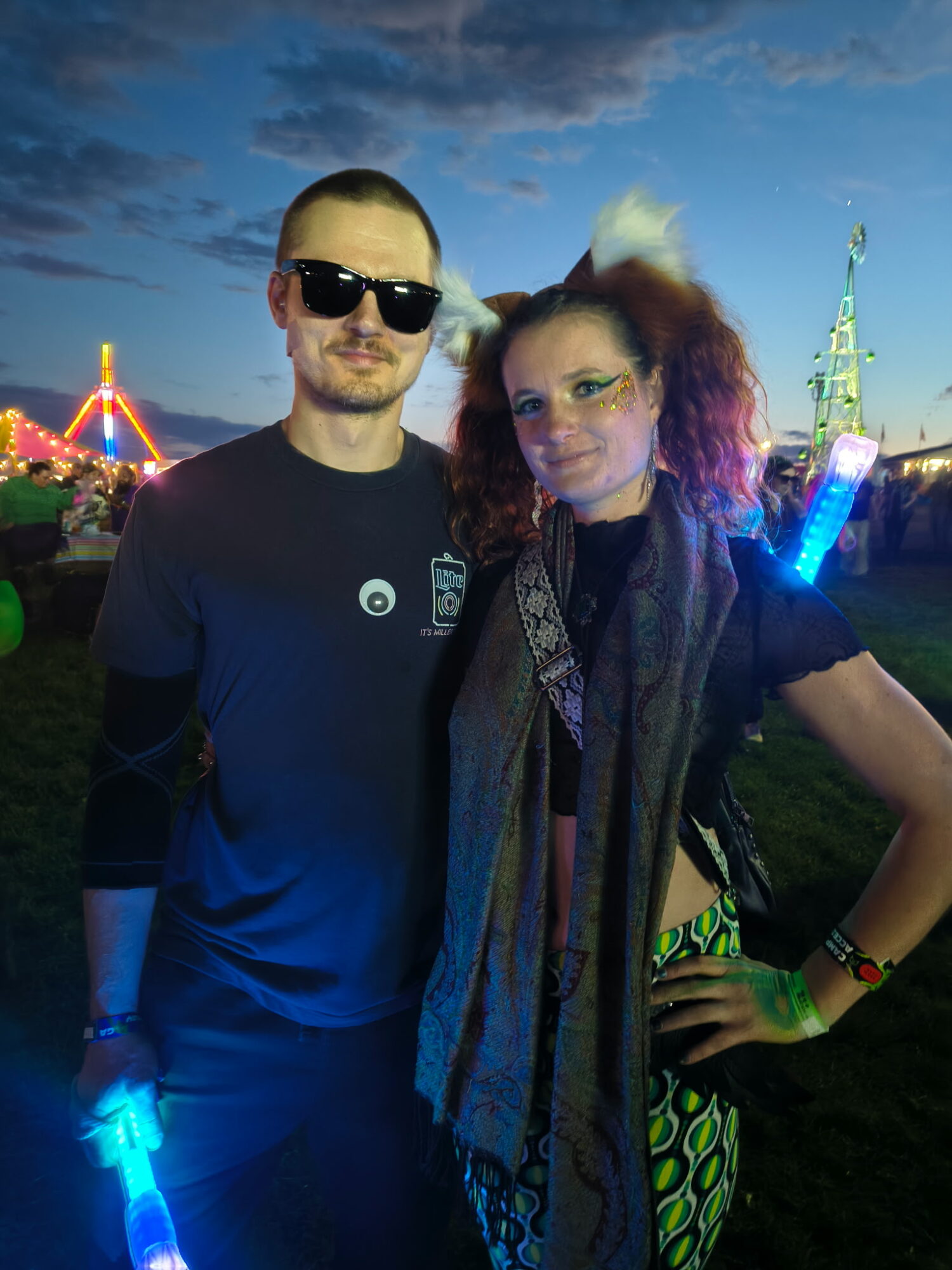 Two people at a nighttime outdoor event, one wearing sunglasses and the other with colorful hair, illuminated by glow sticks.