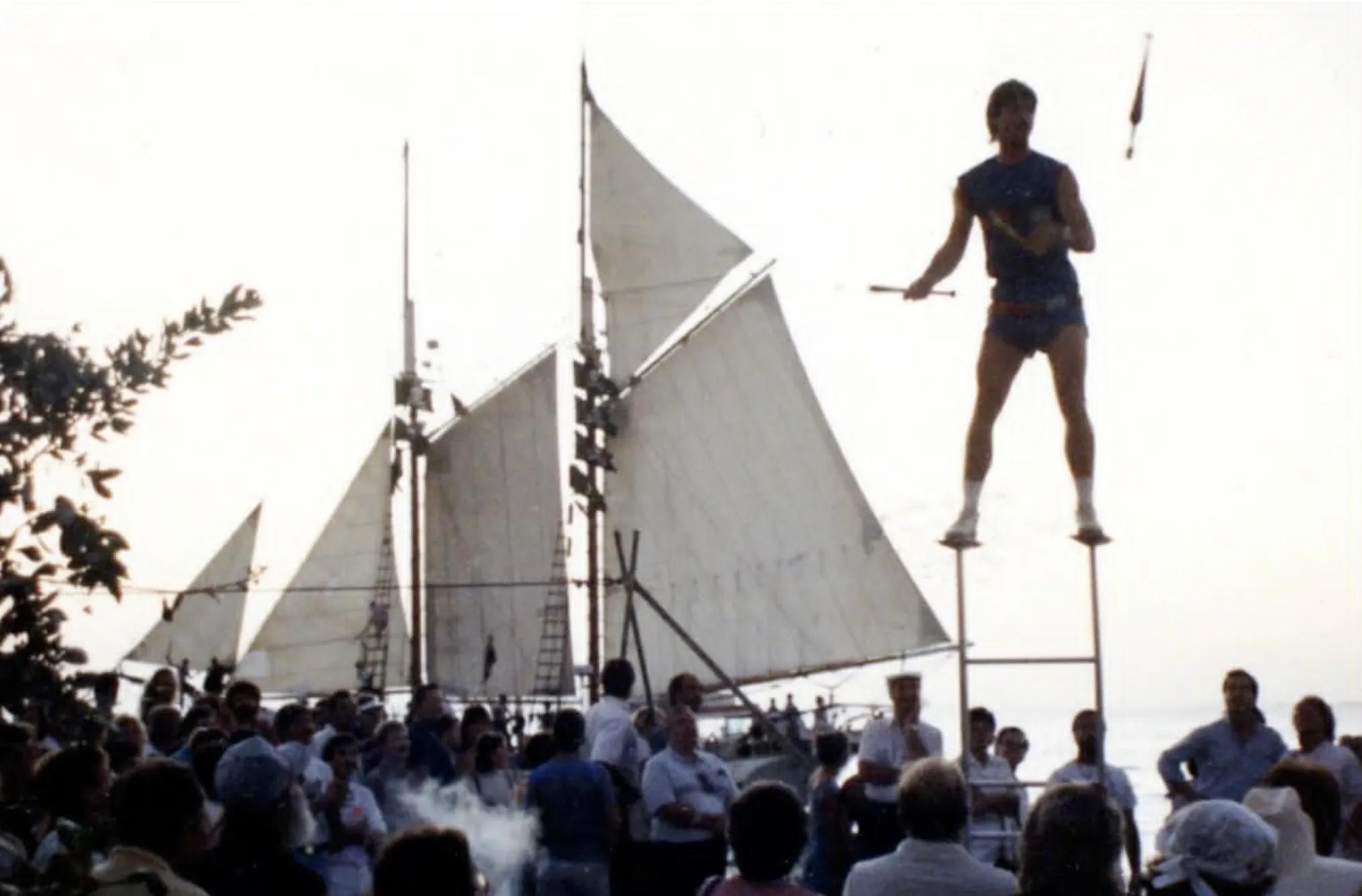 A large crowd watches a performer standing on a tall platform during an outdoor event with sailboats in the background.