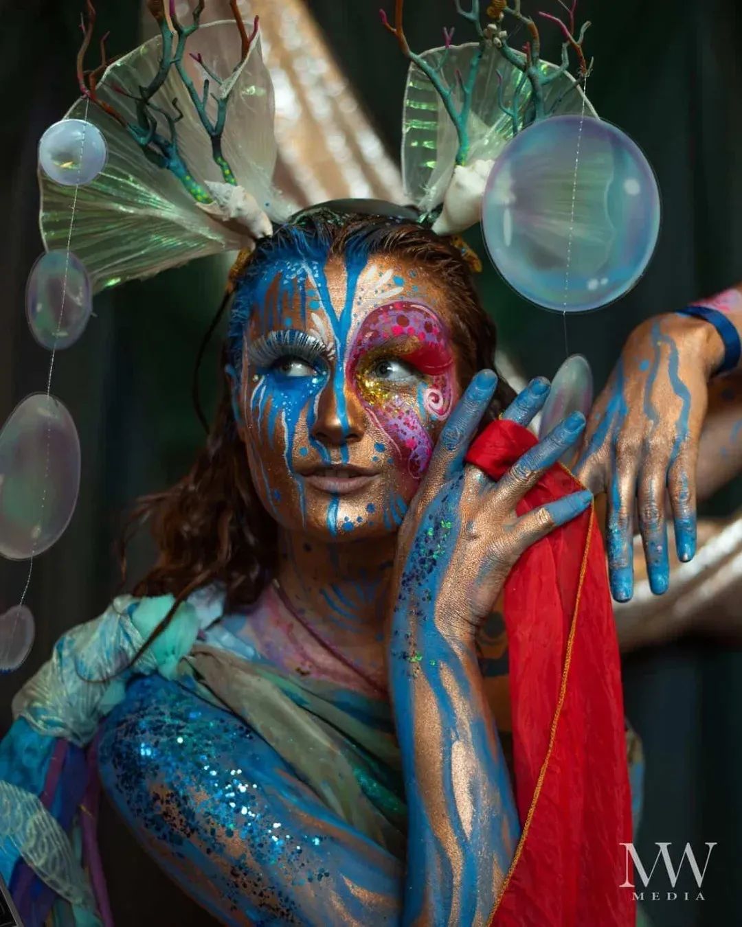 Person with elaborate face paint, wearing headpiece with branches and orbs, holding a red object, colorful makeup and clothing.