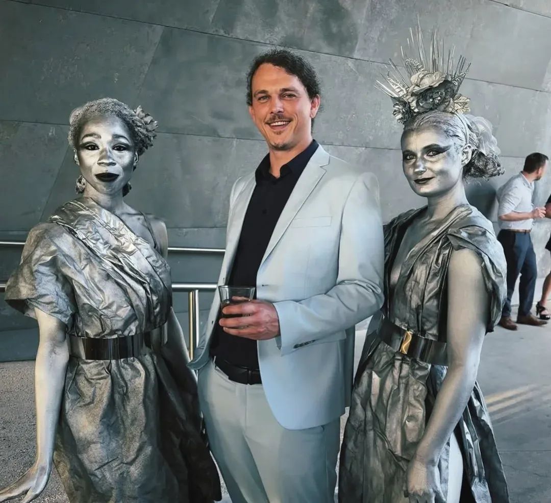 Man in light suit with two performers in metallic costumes and makeup, standing outdoors against a gray wall.