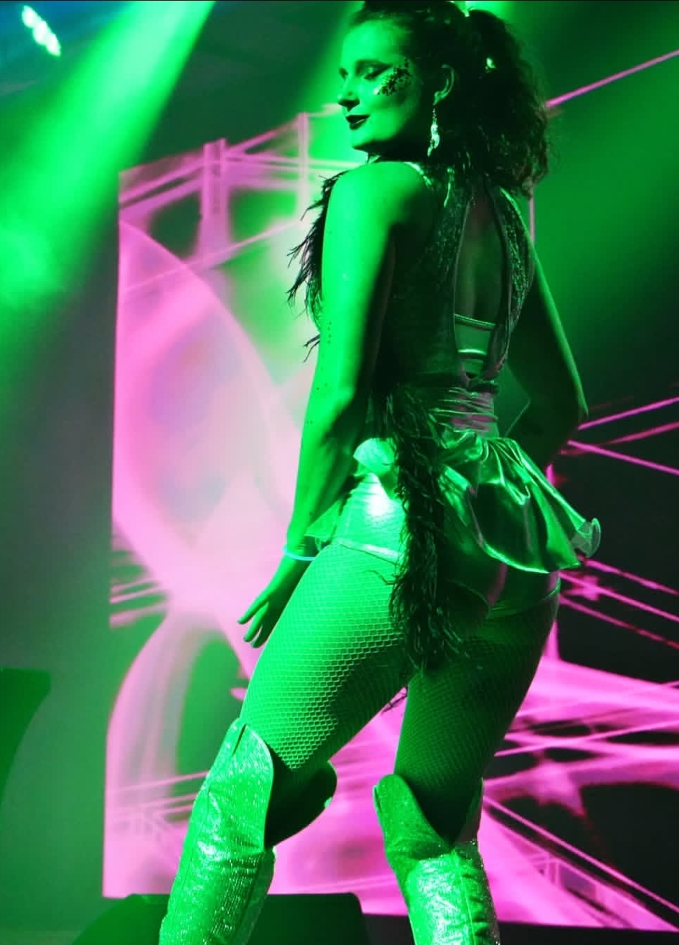 Performer on stage with green and pink lighting, wearing a black outfit and thigh-high boots, striking a pose.