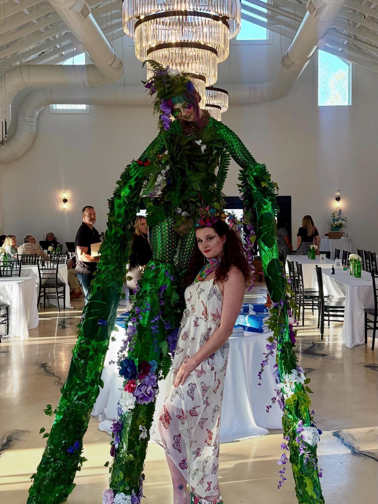 Woman in floral dress standing next to a large green plant sculpture in a decorated event hall.