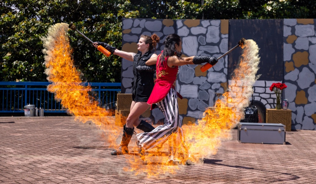 Two performers spray fire in opposite directions, creating arcs of flames on a paved outdoor area with a stone wall background.