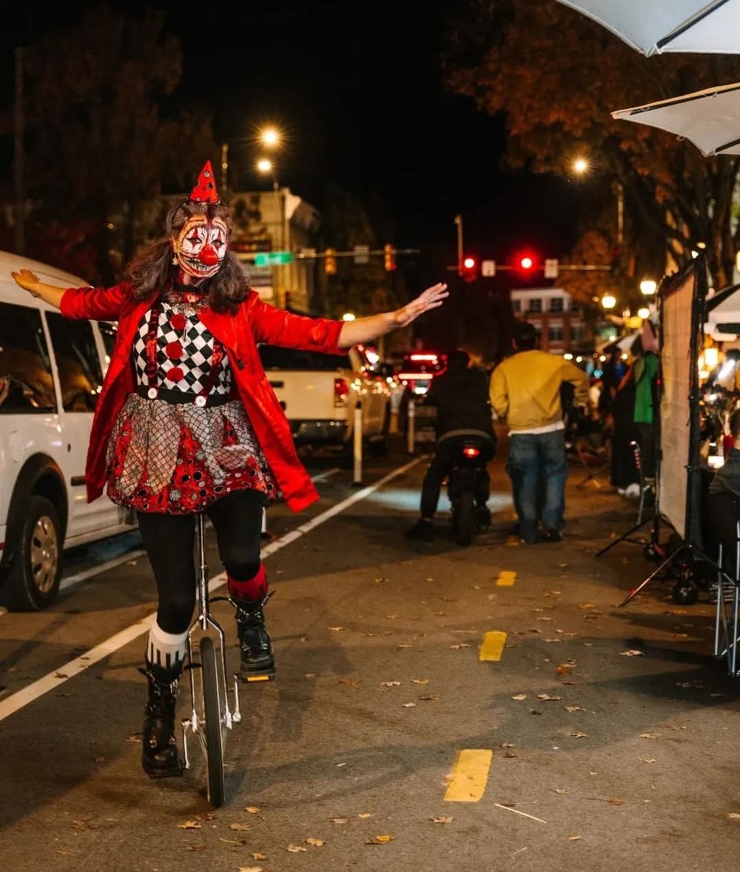Person dressed as a clown balancing on a unicycle on a city street at night, with people and cars nearby.