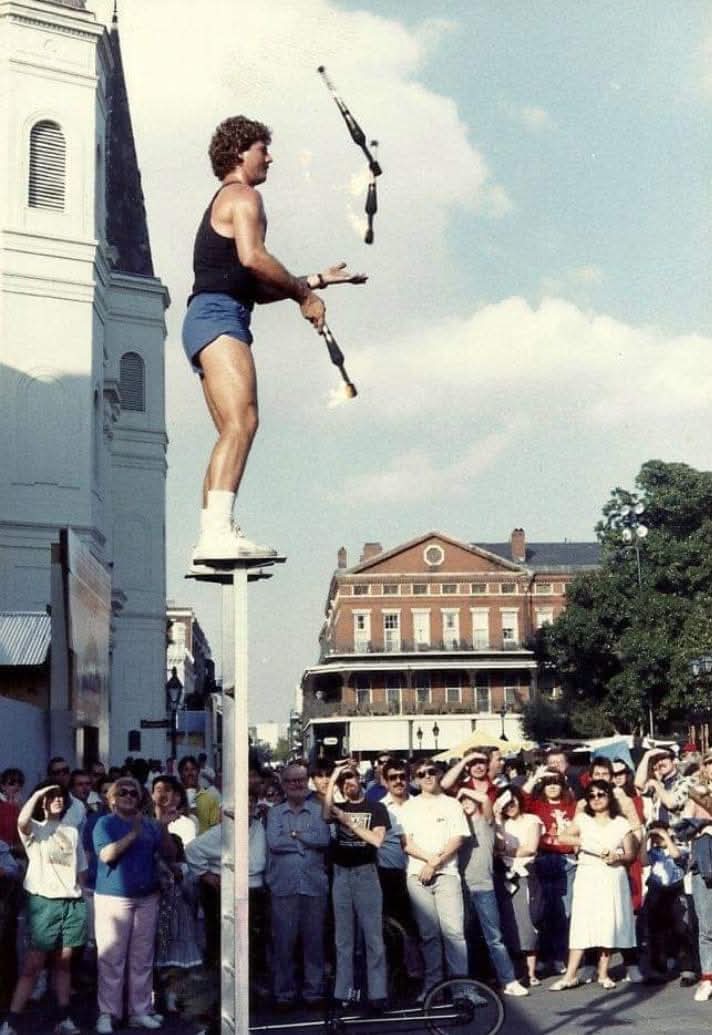 A person balancing on a tightrope while juggling objects above a crowd outdoors, with buildings and trees in the background.