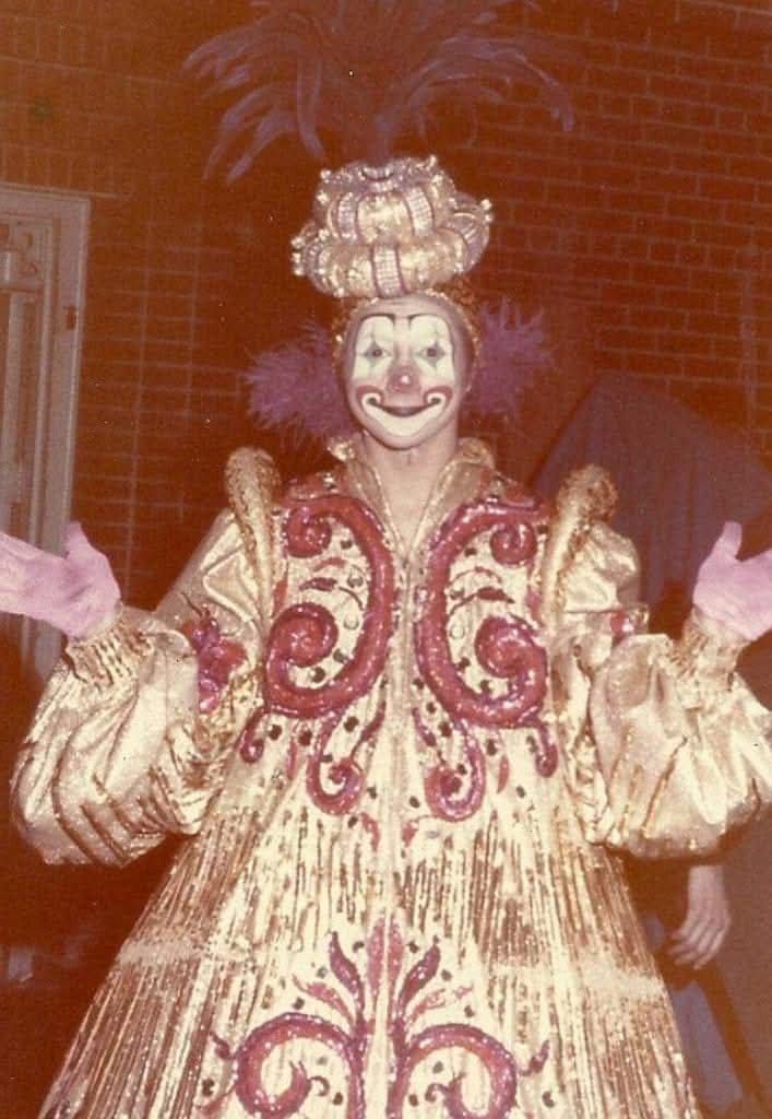 Person dressed as a clown with a large headpiece, colorful costume, and painted face, standing outdoors at night.
