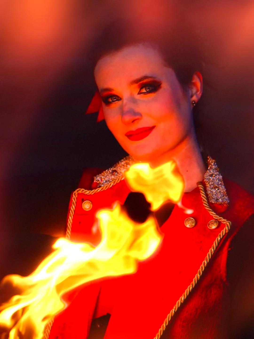 Woman with makeup and earrings, wearing a red jacket with gold details, smiling in a dark background with flames.
