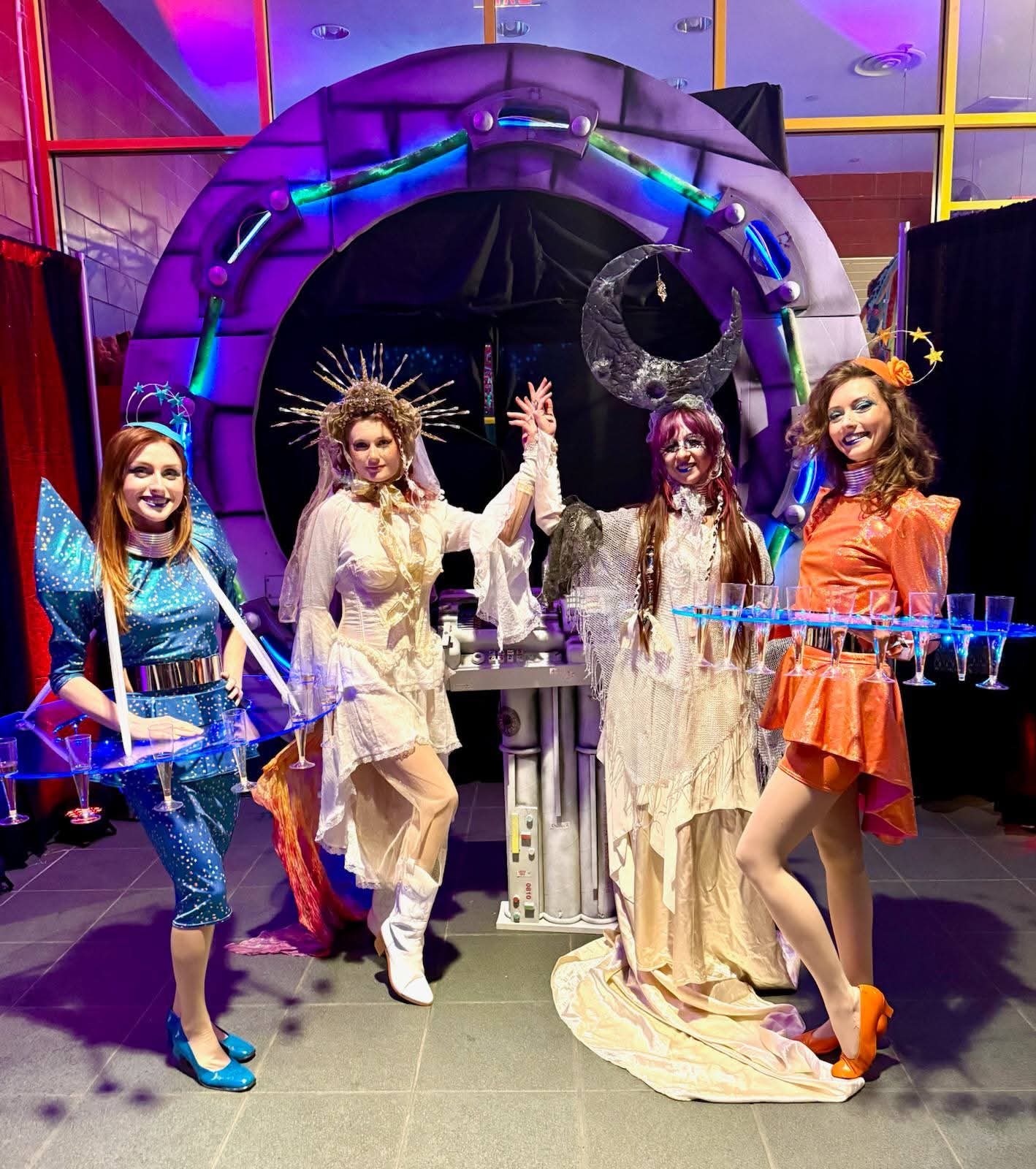 Four women in colorful costumes pose in front of a space-themed backdrop with a moon and planets, holding props. Word count: 20.