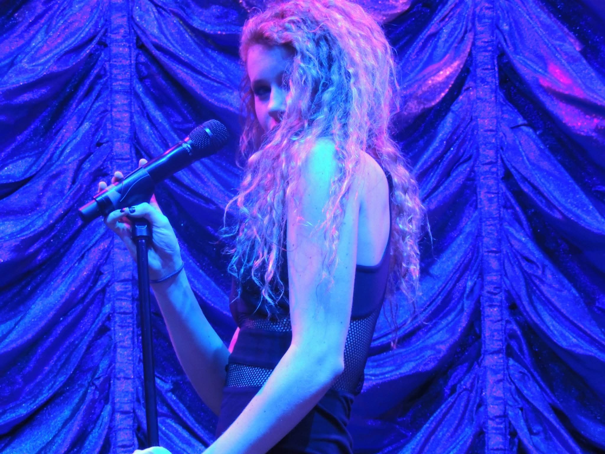 A woman with long, curly hair stands on stage holding a microphone, with a purple curtain backdrop.