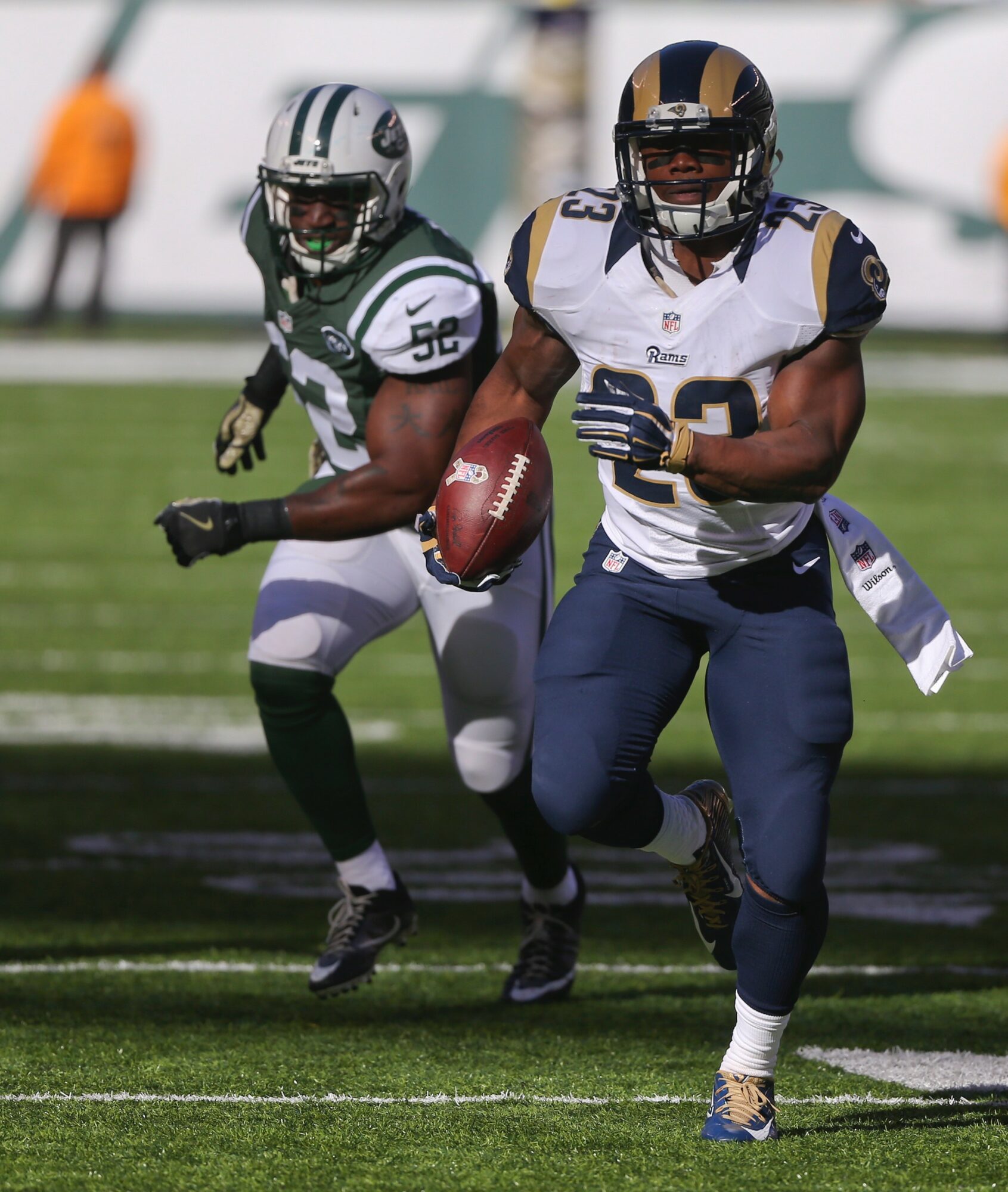 Football player in white and blue uniform running with ball, defender in green and white behind, on field.