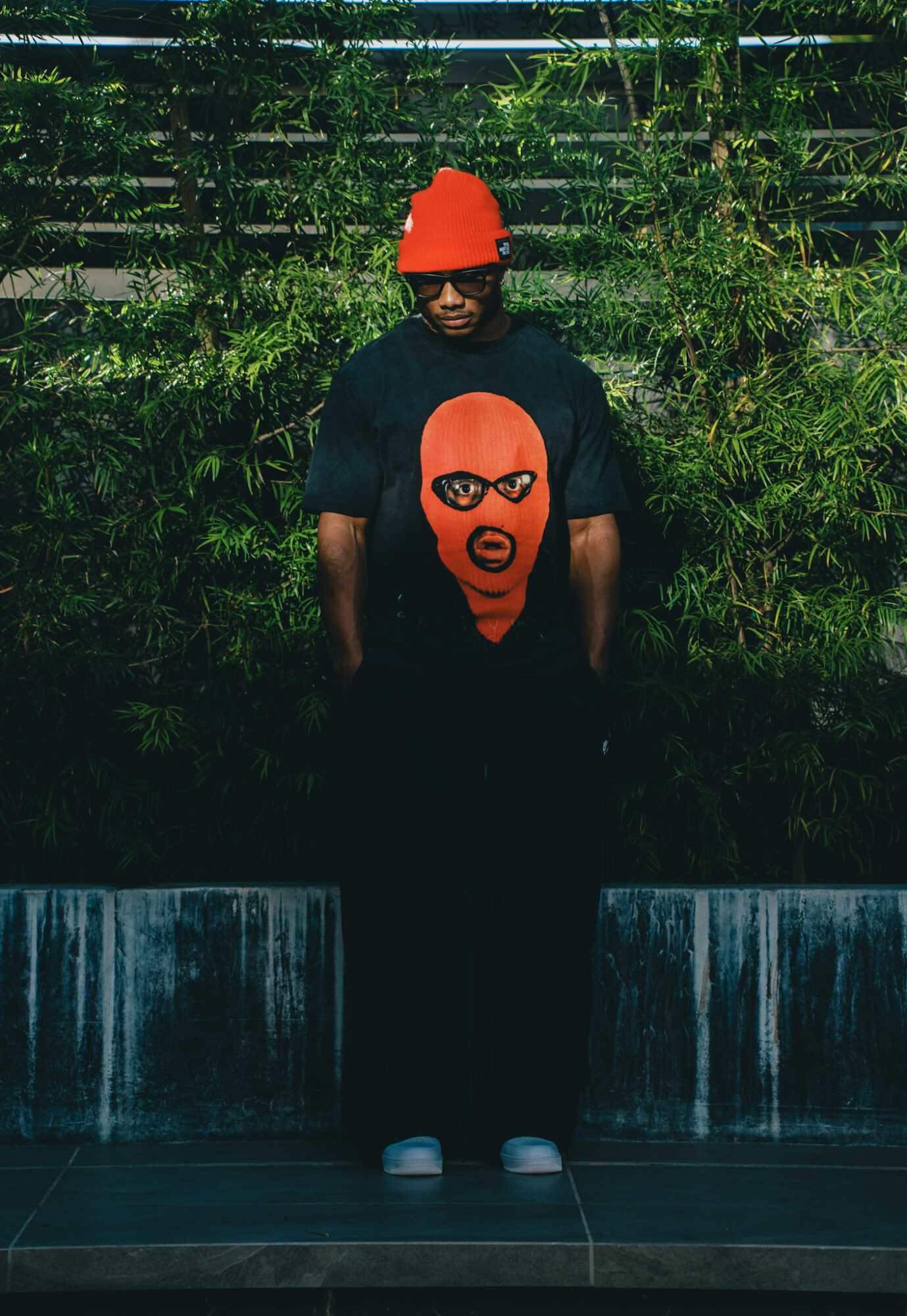 Person wearing a red hat and sunglasses, standing outdoors in front of greenery and a water feature, wearing a black shirt with a red face graphic.