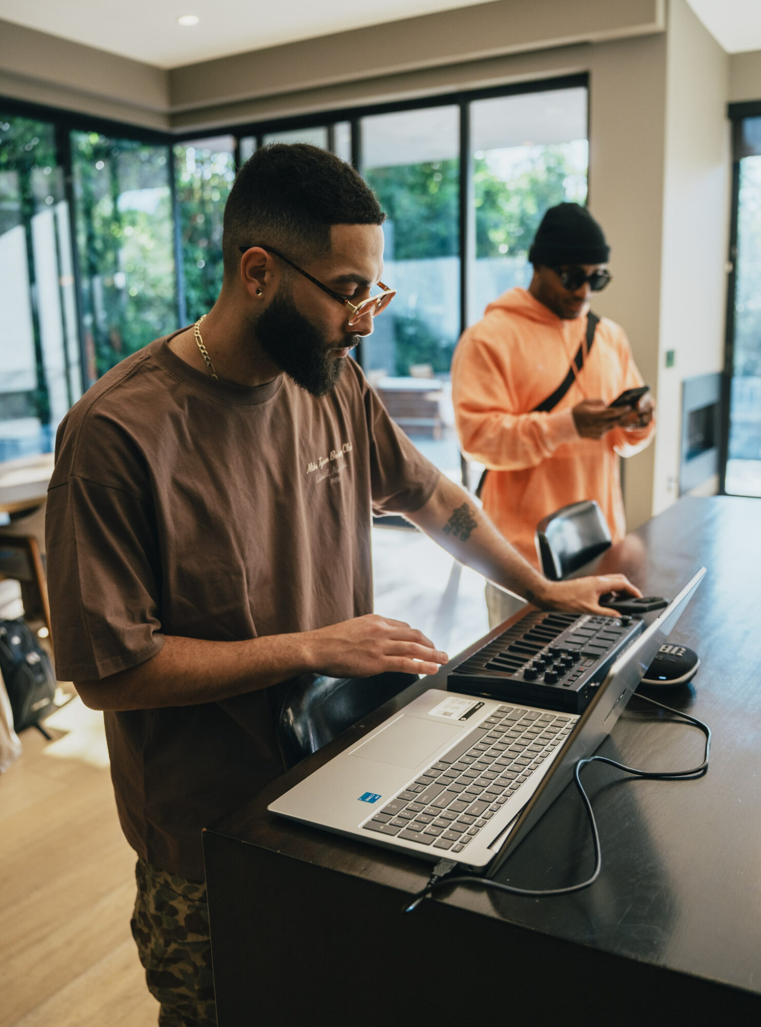 Two men working at a counter with laptops and a phone in a modern room with large windows showing greenery outside.