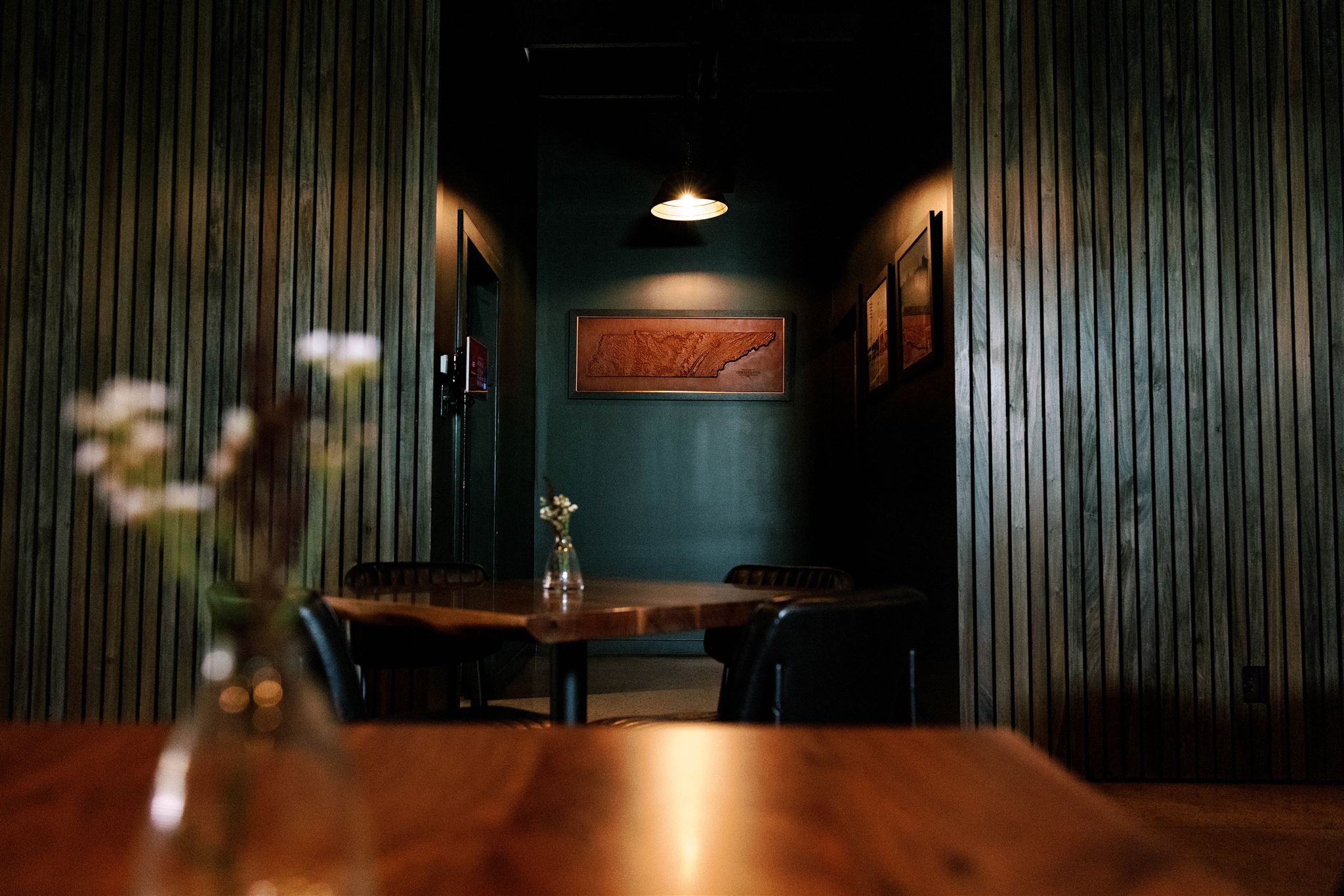 View of a dimly lit room with a table, chairs, and wall art, framed by wooden-paneled walls.