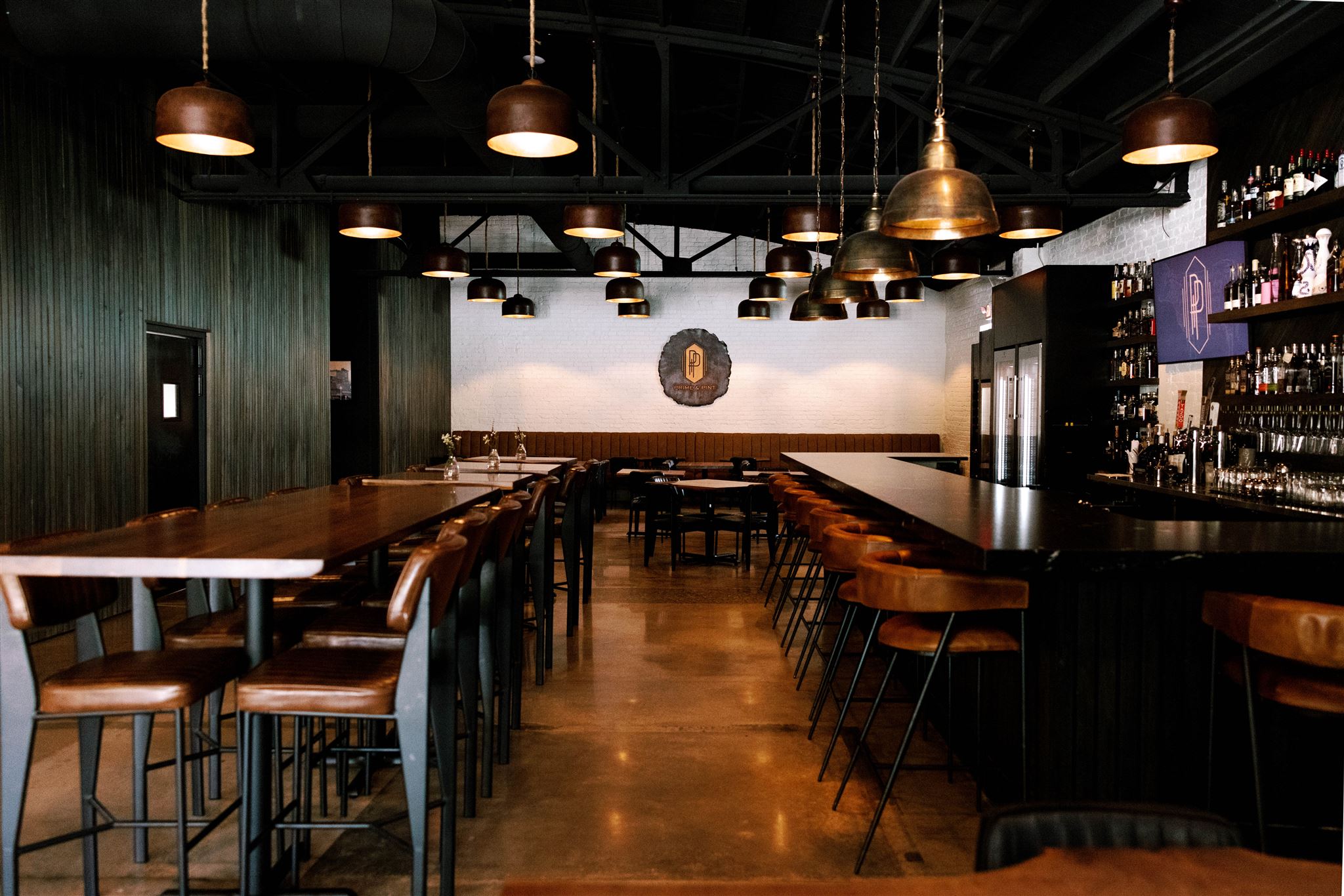 Bar with wooden tables and chairs, pendant lights, and a bar counter with bottles, against a wall with a clock.