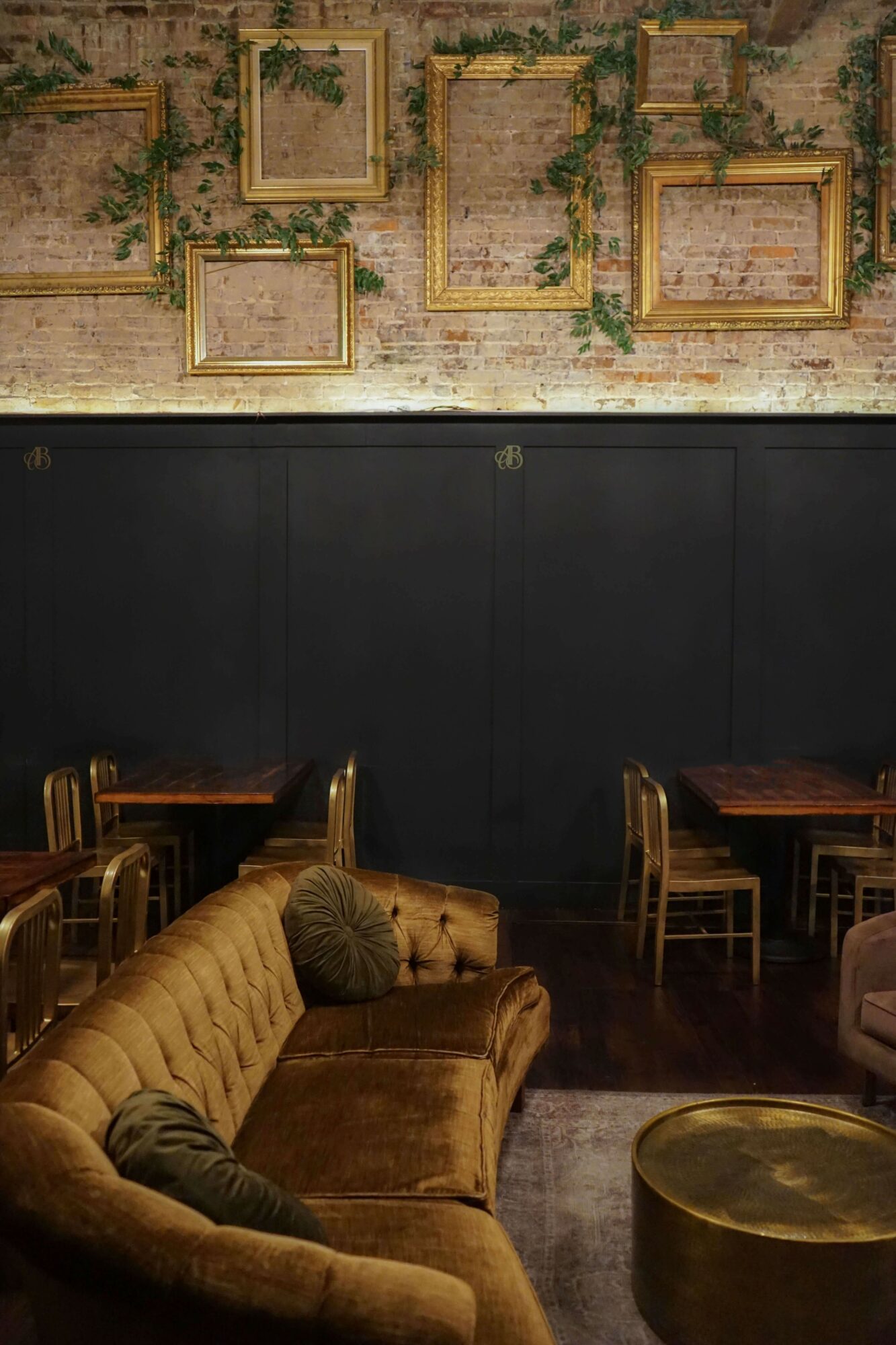 Interior of a cafe with a mustard-colored sofa, wooden chairs, framed artwork on a brick wall, and a round table.