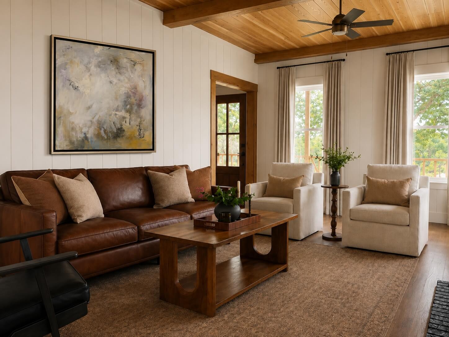 Living room with a brown leather sofa, two white armchairs, a wooden coffee table, and large windows with curtains.