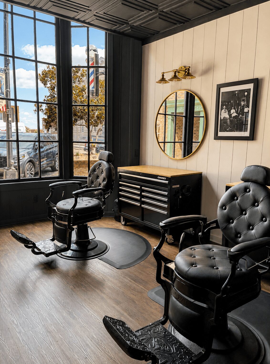 Interior with large window, black barber chairs, round mirror, framed photo, and wooden cabinet, sunlight streaming in.
