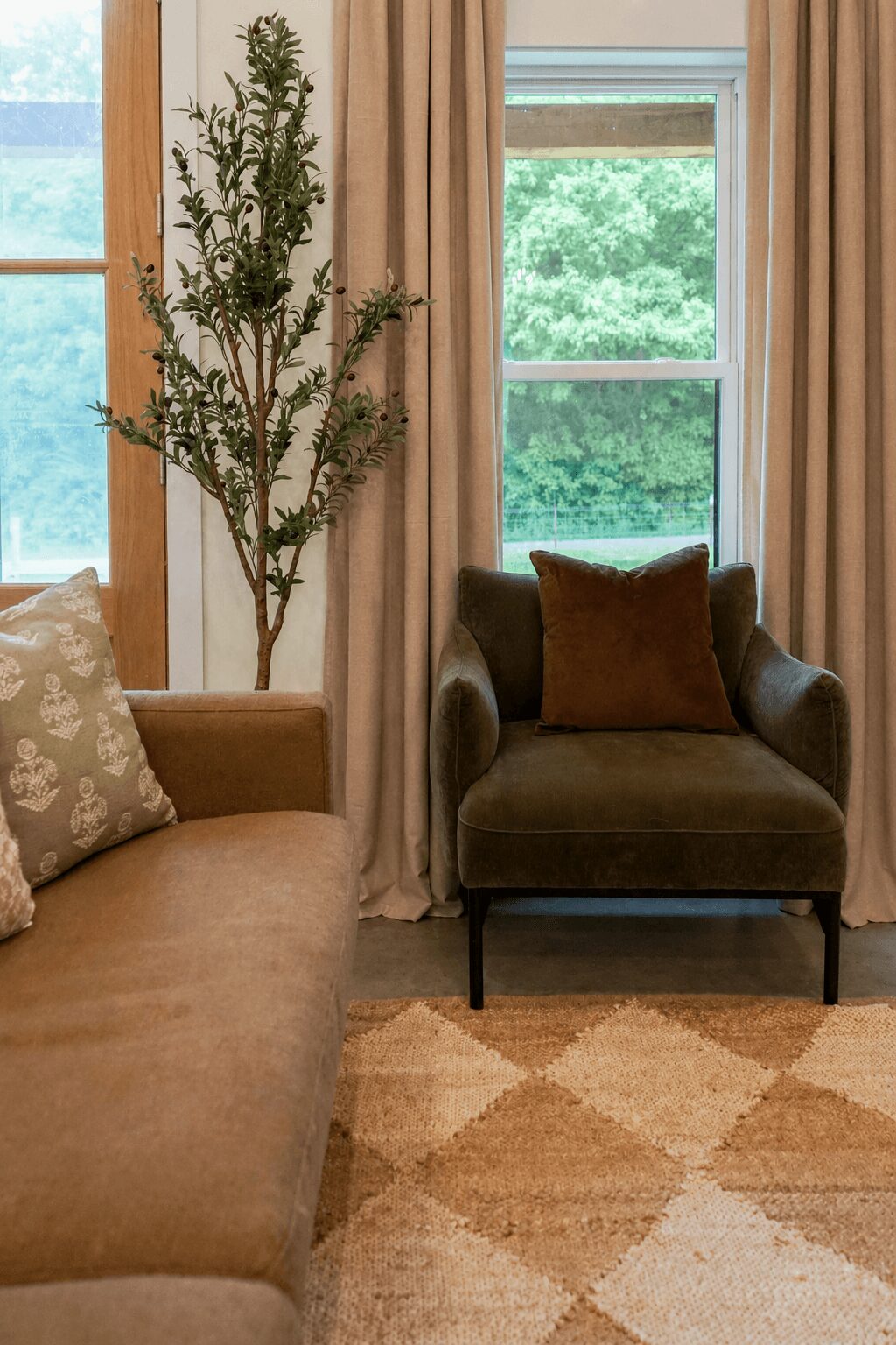 Living room with a brown sofa, a dark armchair with a pillow, a tall plant, and windows with curtains, greenery outside.
