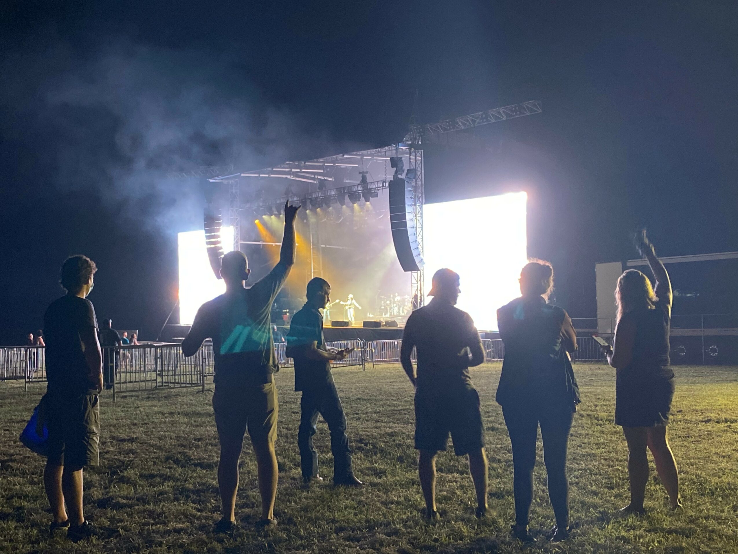 Group of six people watching a concert stage with bright lights and smoke, some raising hands, outdoors at night.