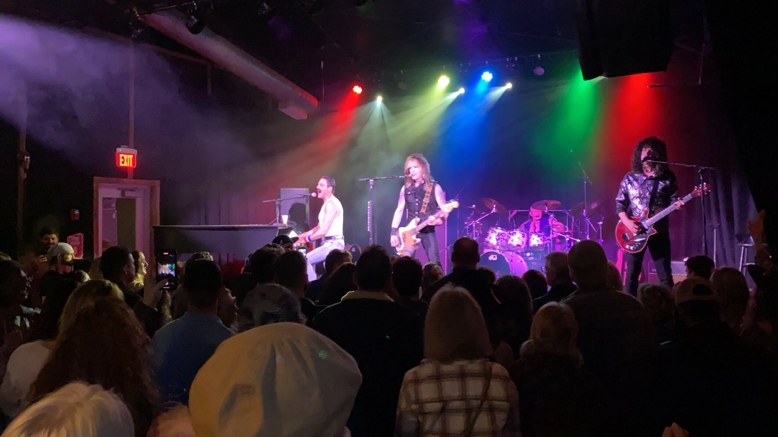 Band performs on stage with colorful lights, audience watching in a dark venue, some people with hats, some with long hair.