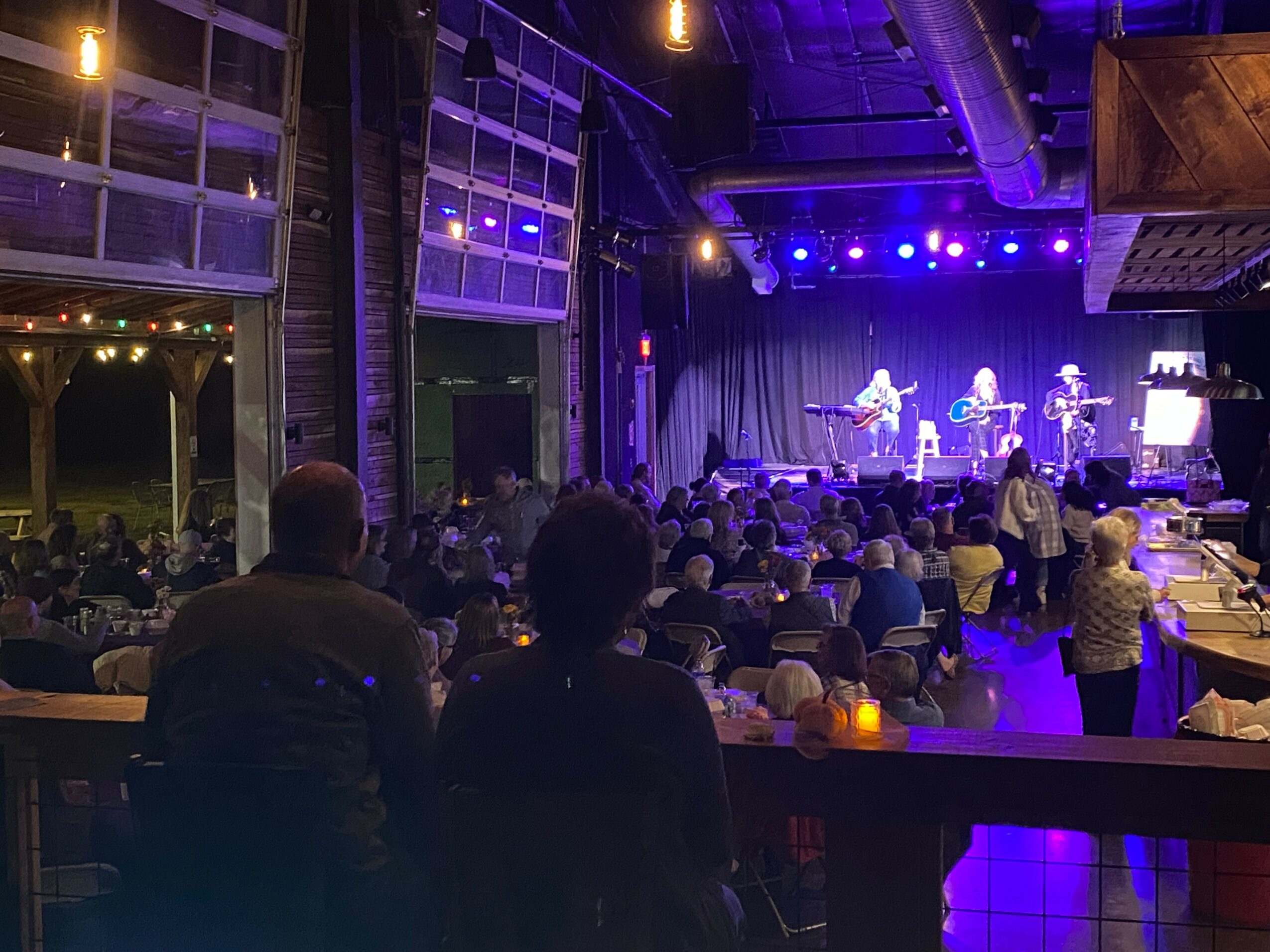 Audience watching a live band perform on stage in a dimly lit venue with purple and blue lighting.