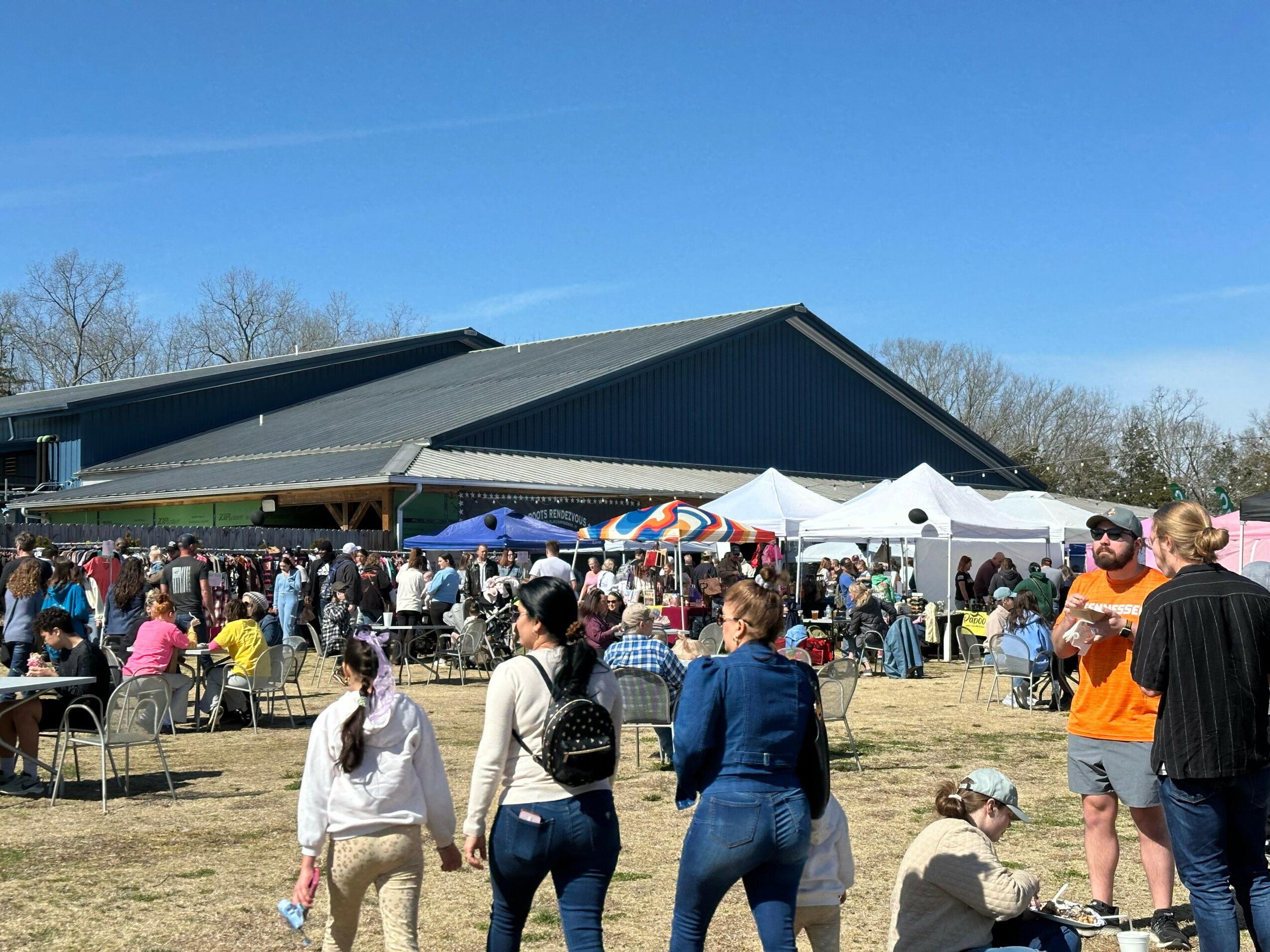 People gather outdoors near a large building with tents and umbrellas, some sitting and others walking, under a clear blue sky.