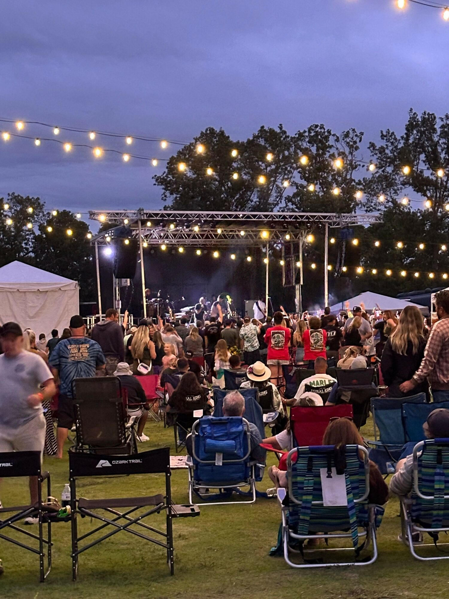 People gathered outdoors at night with string lights and trees, seated on chairs, attending an event or concert.