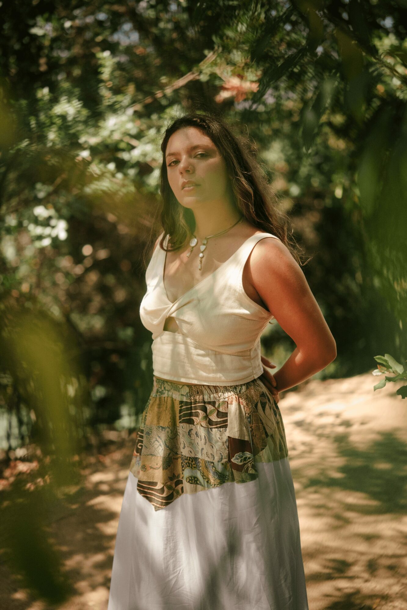 Young woman with long dark hair standing outdoors among trees, wearing a white sleeveless top and patterned skirt, sunlight filtering through leaves.