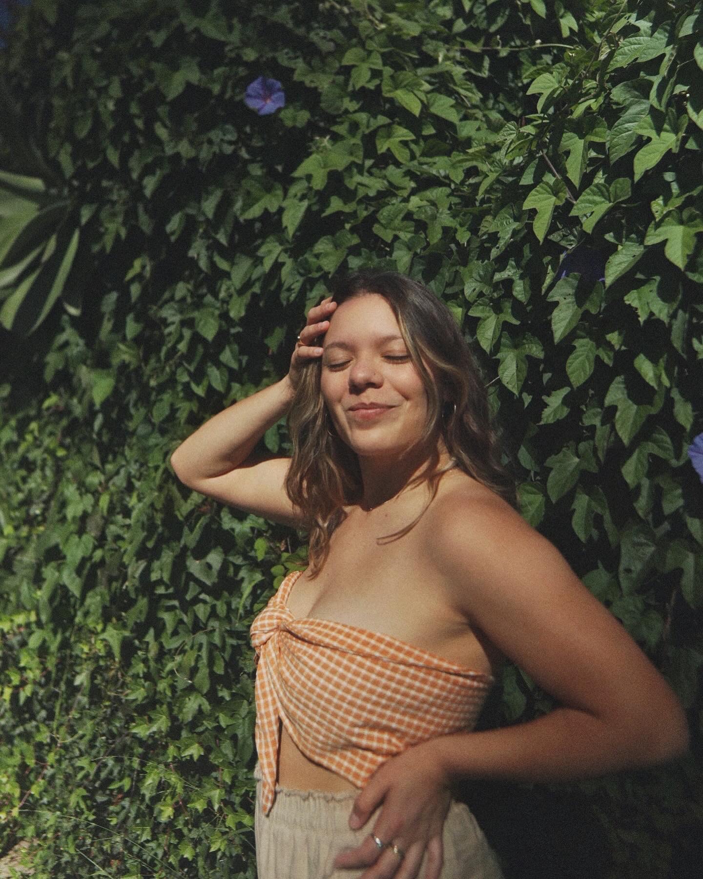Young woman with long wavy hair smiling with eyes closed, standing outdoors against green leafy background, wearing a strapless orange checkered top.