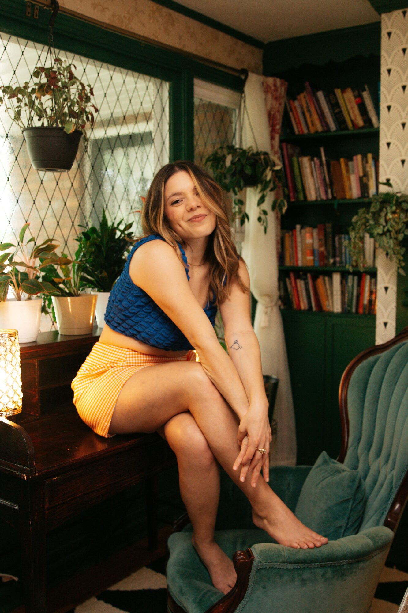 Young woman with long hair sitting on a windowsill in a cozy room, smiling, surrounded by plants and books.
