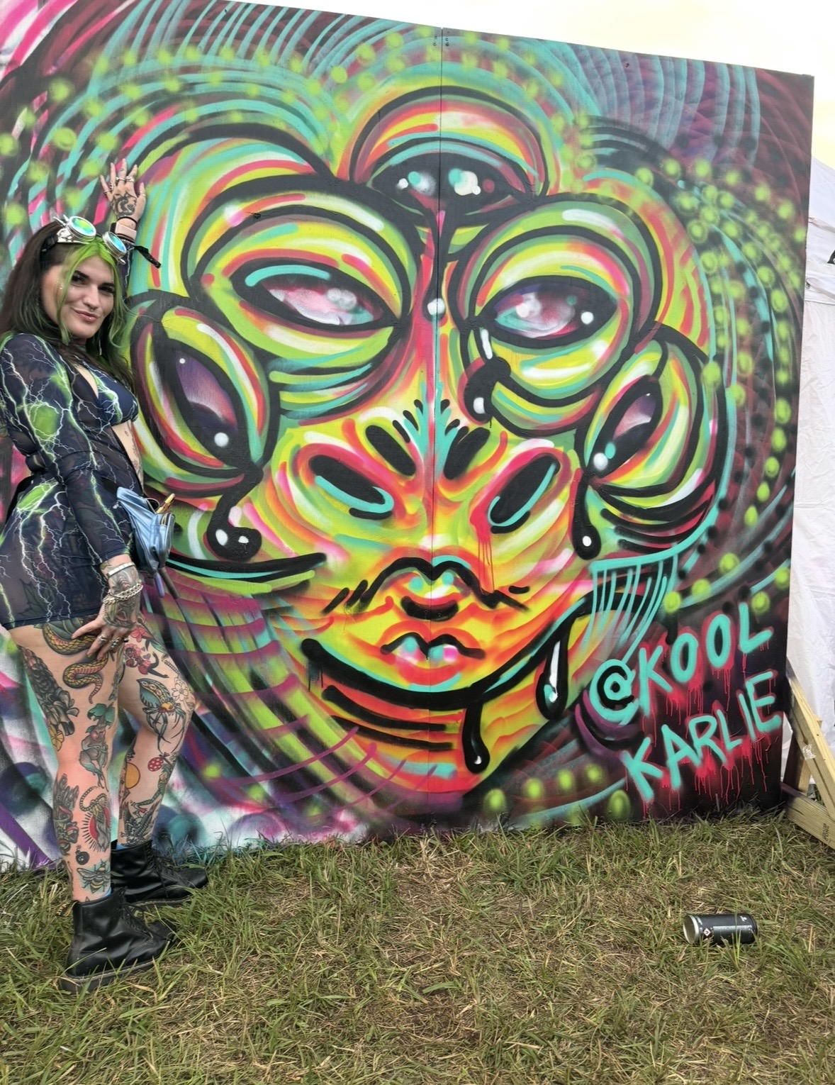 Woman standing next to a colorful graffiti mural of a face with multiple eyes and abstract features, with the artist's signature 'KOOL KARLE'.