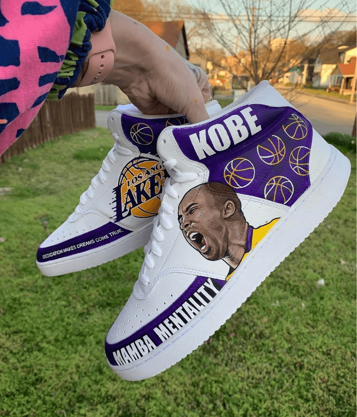White sneakers with Kobe Bryant's image and Lakers logo, outdoors on grass, with text about Mamba Mentality.