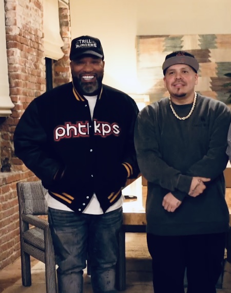 Two men standing indoors, one wearing a black jacket and cap, the other in a dark sweater and cap, smiling.