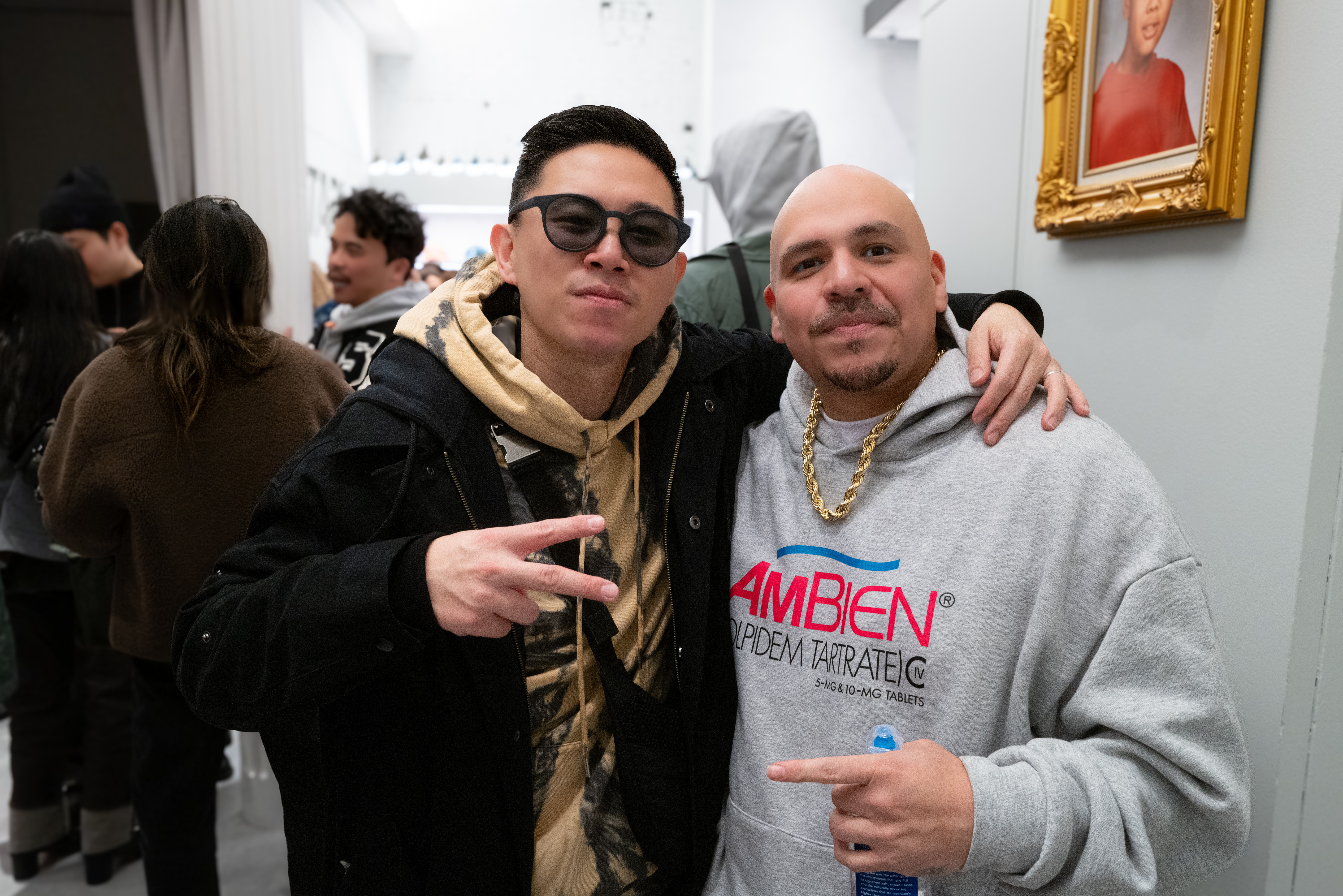 Two men pose together indoors, one wearing sunglasses and a black jacket, the other in a gray hoodie with gold chains, arm around each other.