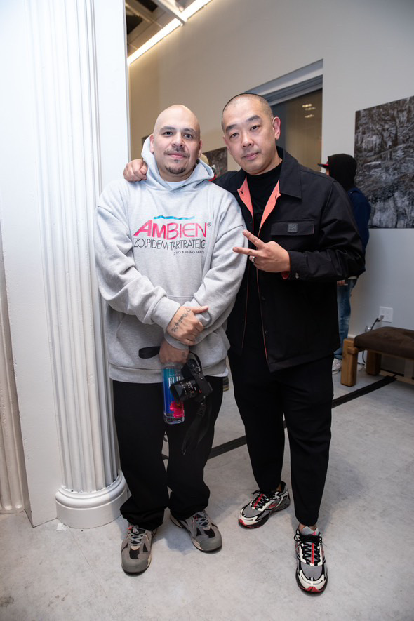 Two men standing close together, one with arm around the other, in an indoor setting with a white wall and a chair in the background.