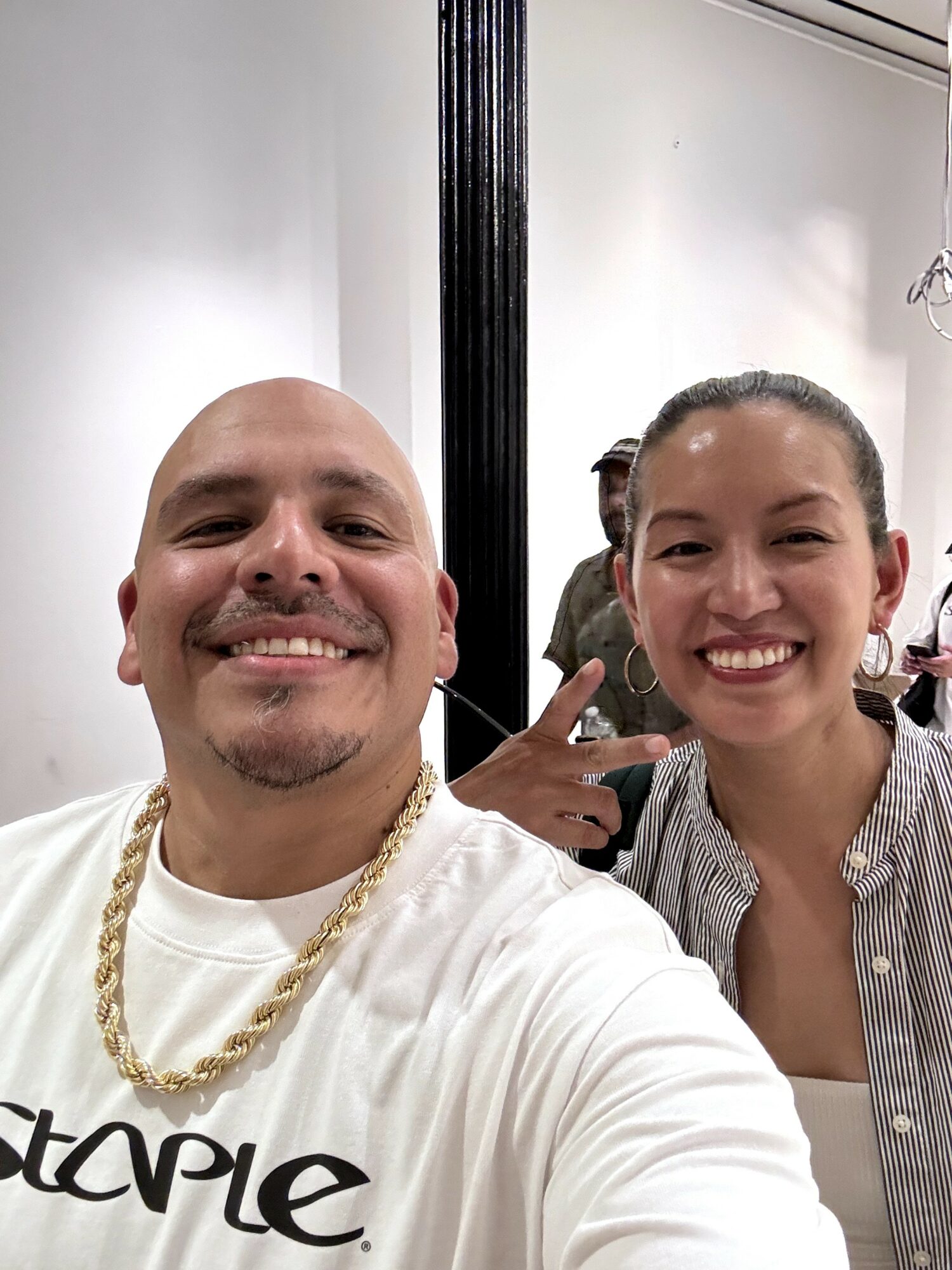 Two smiling people taking a selfie indoors, with a white wall and black vertical beam in the background.