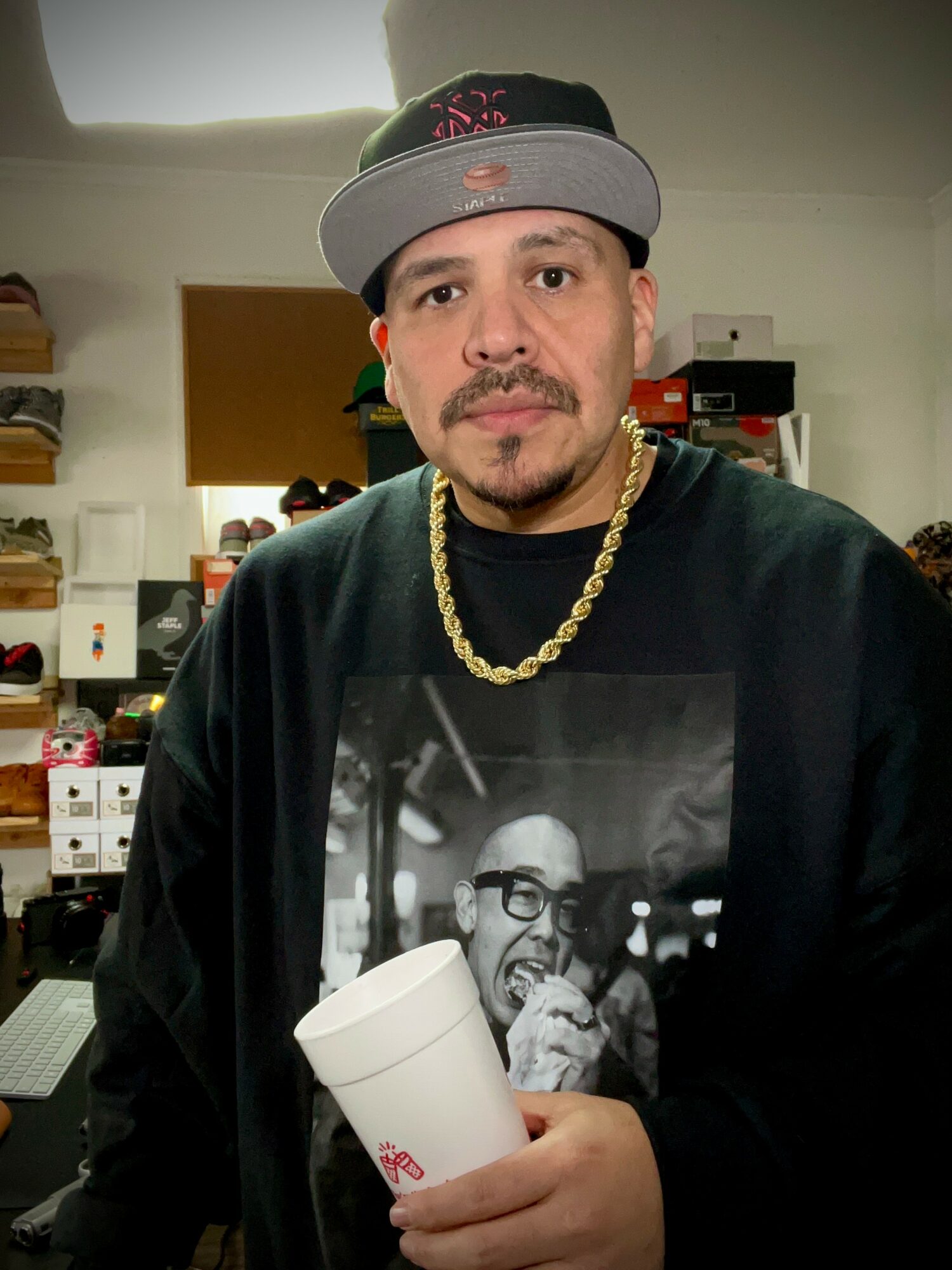 Man wearing a cap and gold chain holding a white cup, standing in a room with shelves and electronic equipment.