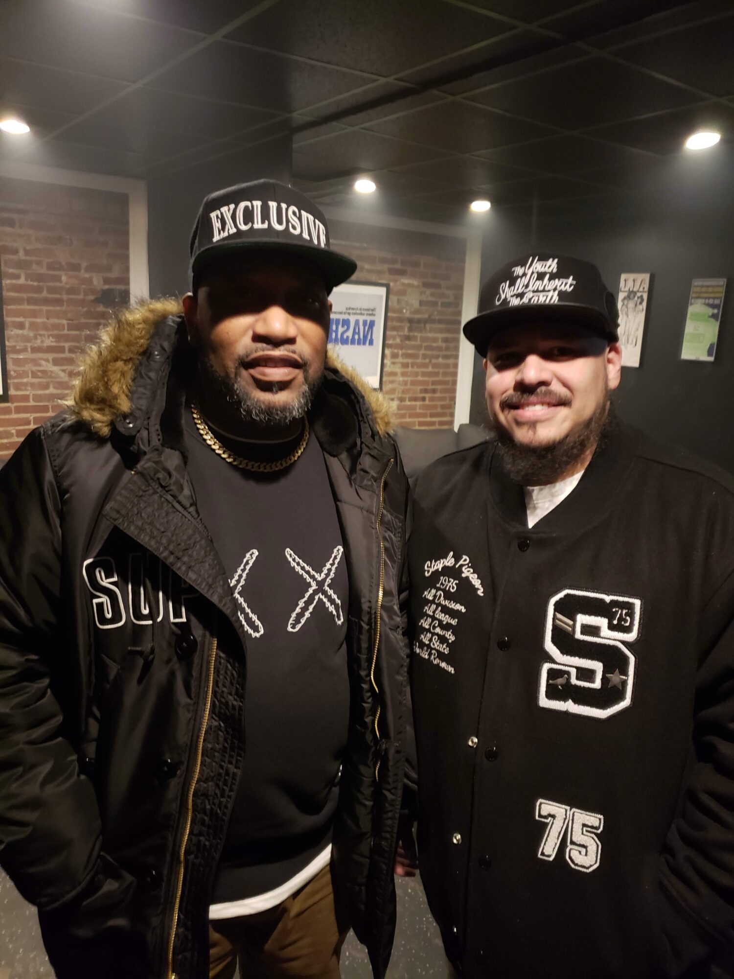 Two men standing indoors, wearing black caps and jackets, smiling at the camera.