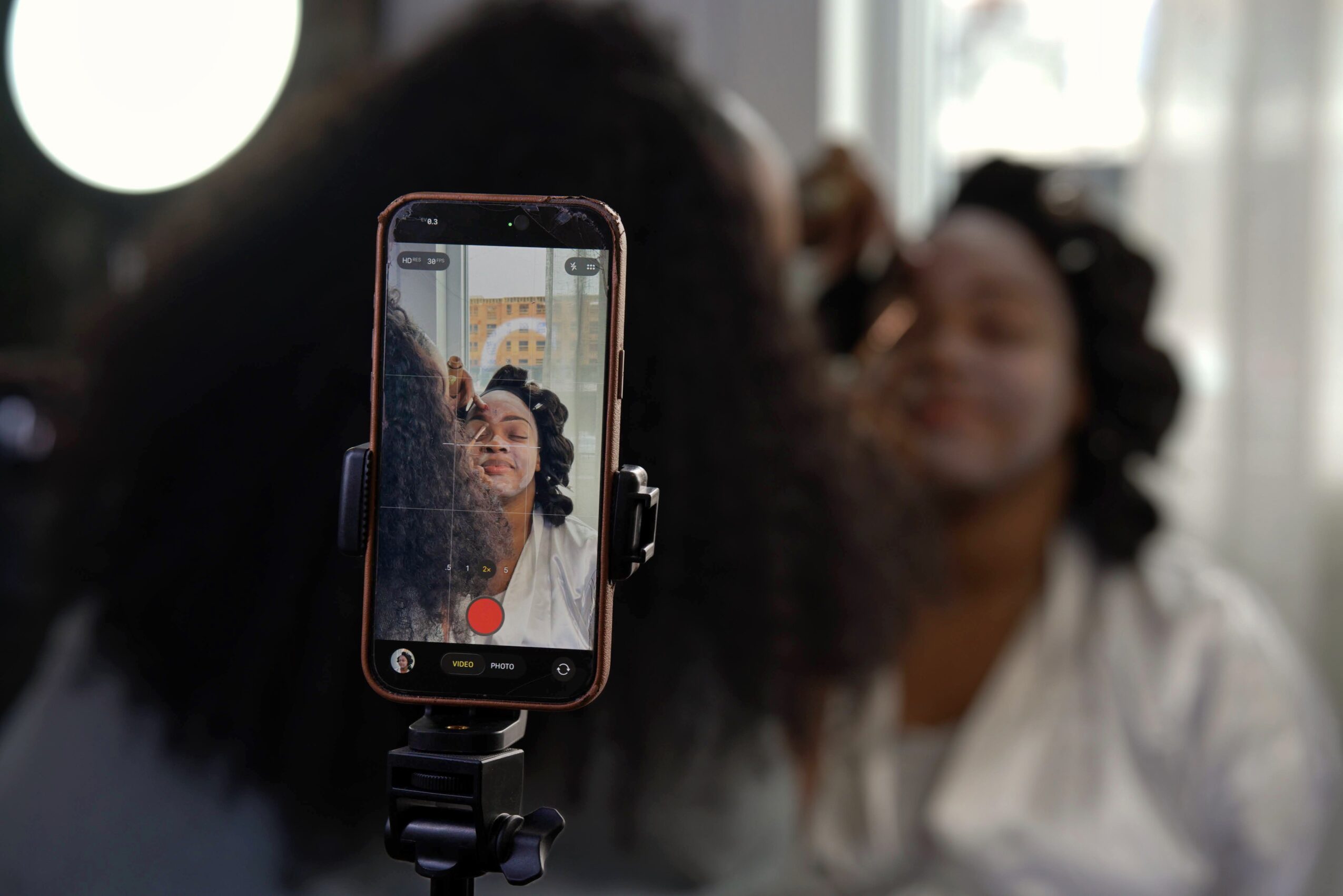 Person taking a selfie with a smartphone, with another person smiling in the background, indoors with natural light.