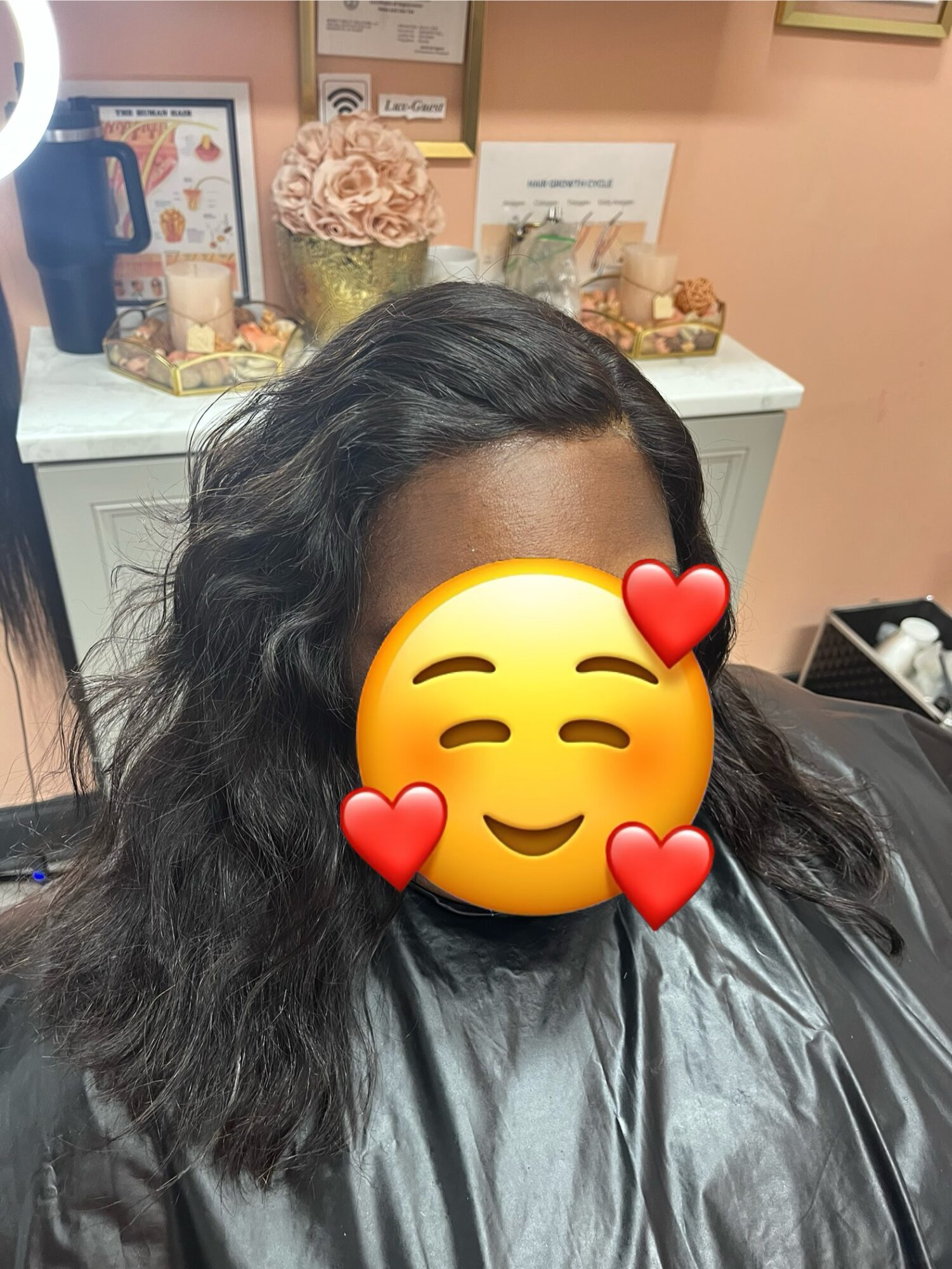 Person with long, wavy black hair sitting in a salon chair, face covered with a smiling emoji with hearts, wearing a cape.