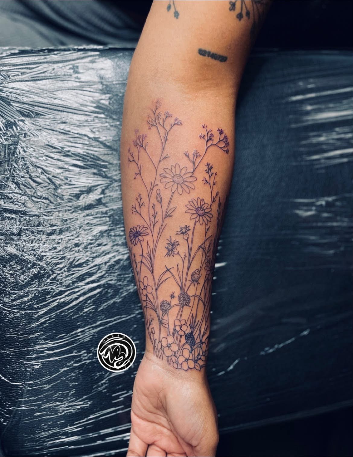 Forearm tattoo of delicate flowers and plants, with a small tattoo above it, against a dark background.