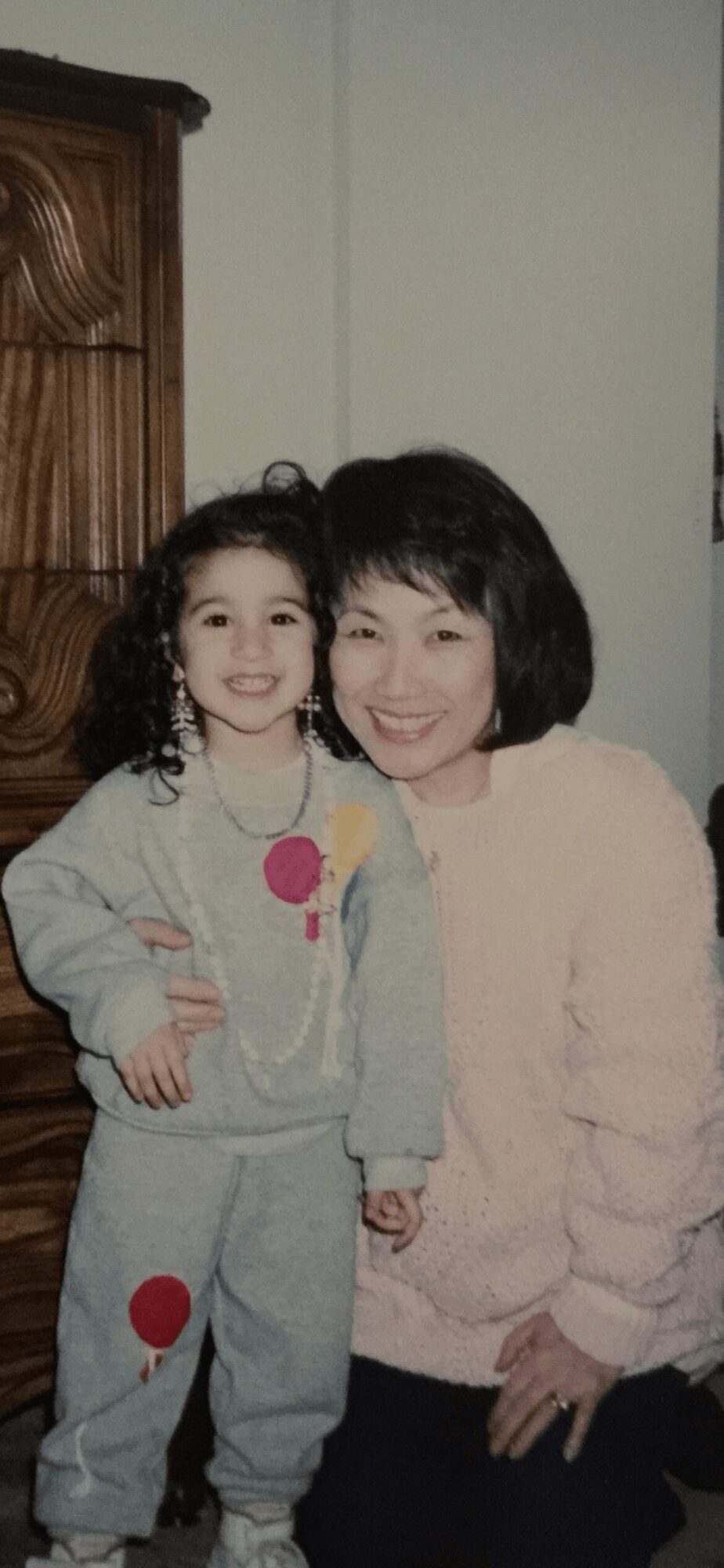 A woman and a young girl stand together indoors, smiling, with a wooden cabinet in the background.