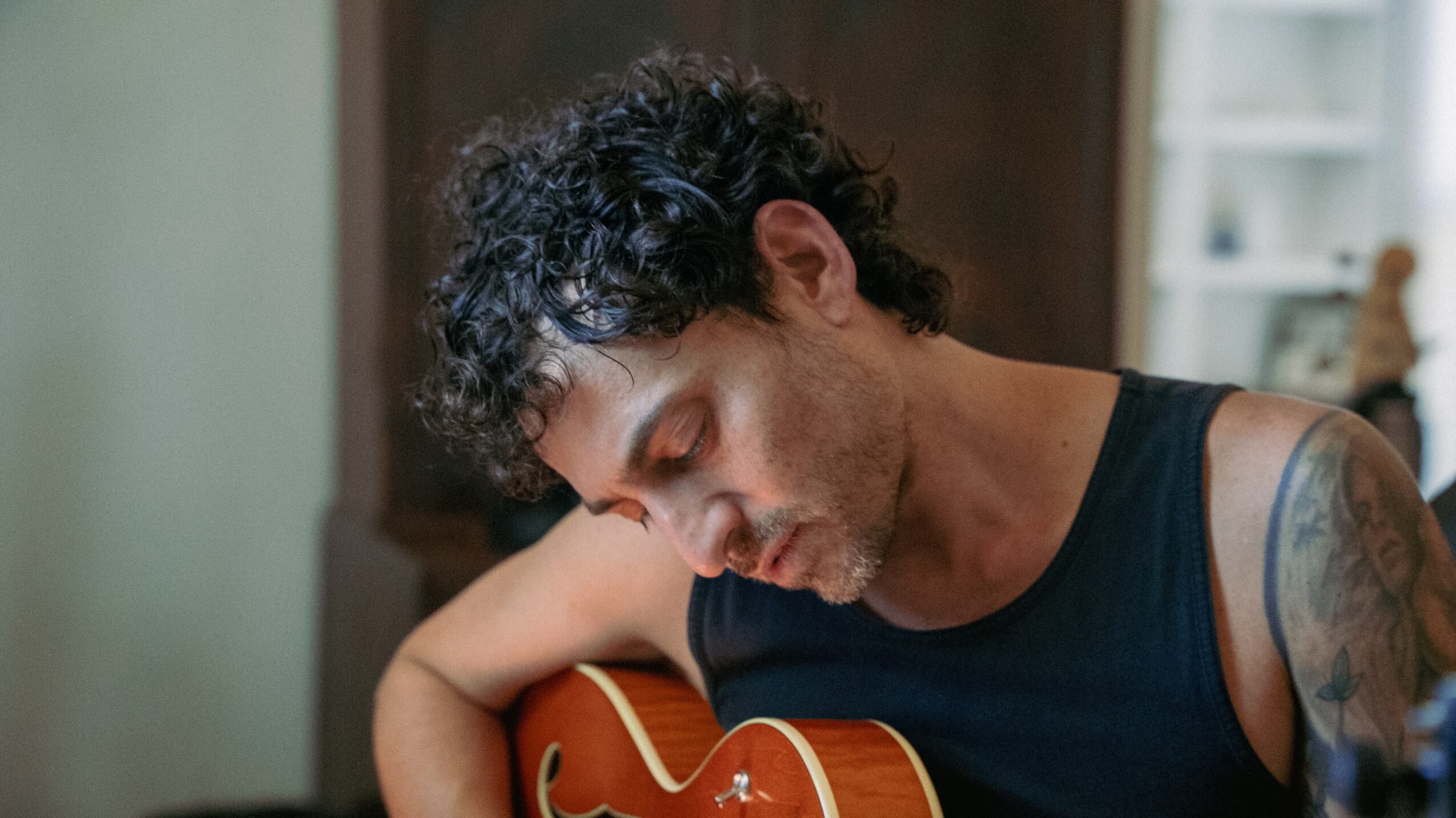 Young man with curly hair playing guitar, focused on his music, wearing a sleeveless shirt, indoors.