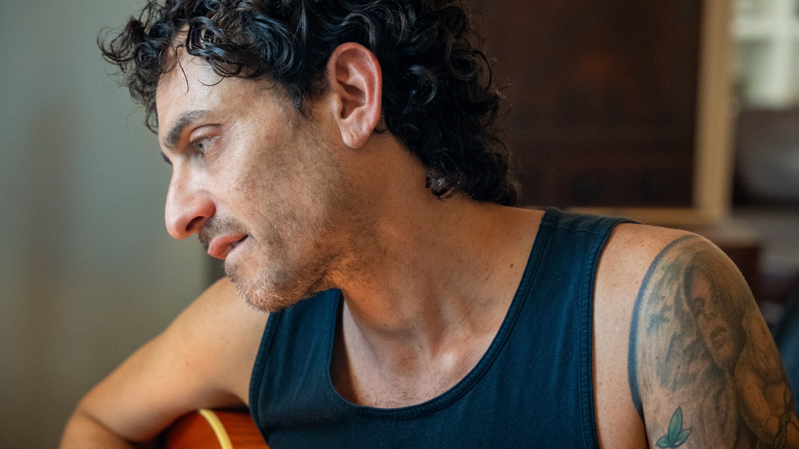 Close-up of a man with curly dark hair, wearing a sleeveless shirt, looking down, with a tattoo on his arm.