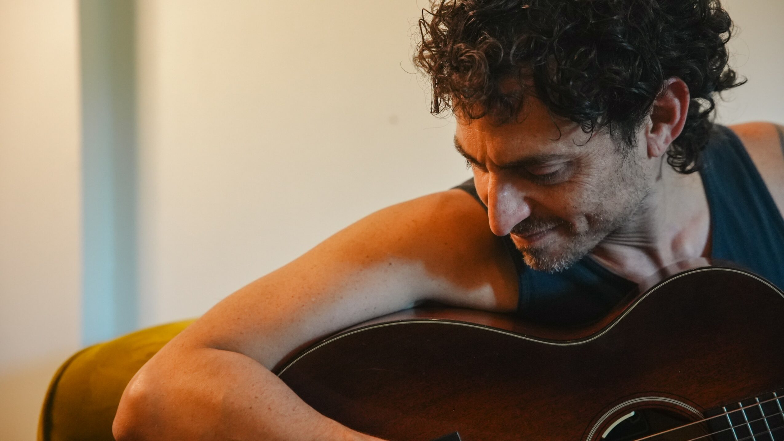 Man with curly hair playing guitar, looking down, in a room with soft lighting.